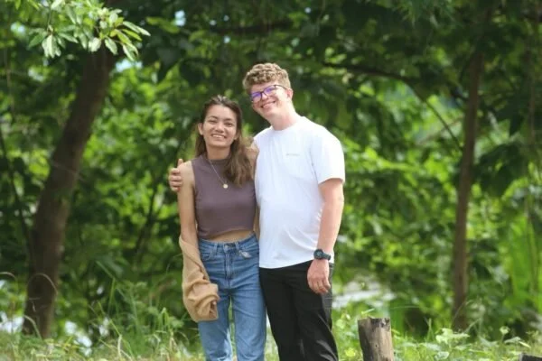 A young woman and a young man smiling and standing outdoors in a green, wooded area.