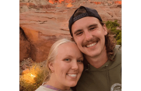 A smiling couple taking a selfie outdoors with a red rock canyon in the background during sunset.