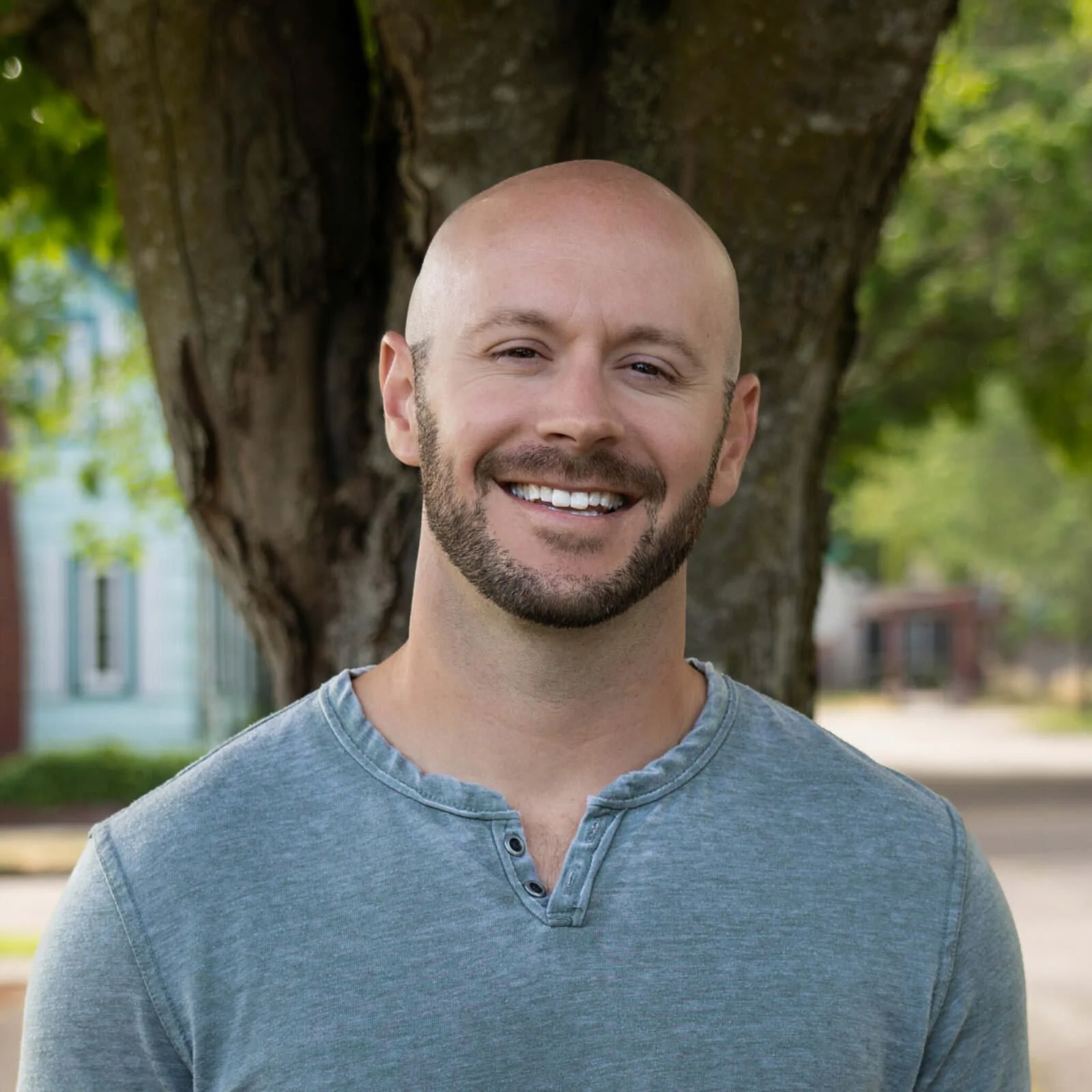 A man with a shaved head and beard smiling outdoors near a tree.