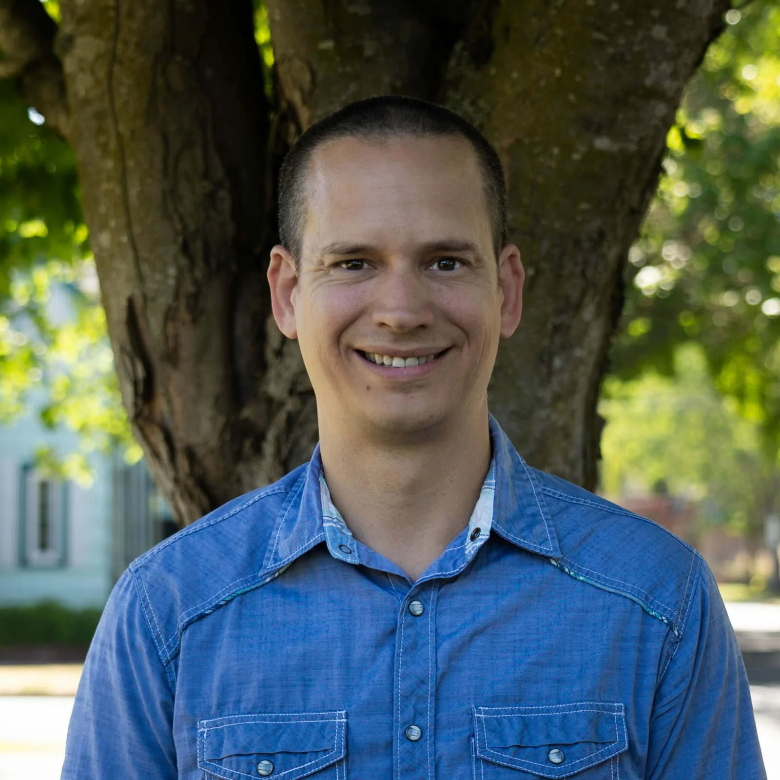 A smiling man with short dark hair, wearing a blue button-up shirt, standing outdoors in front of a tree with green leaves.