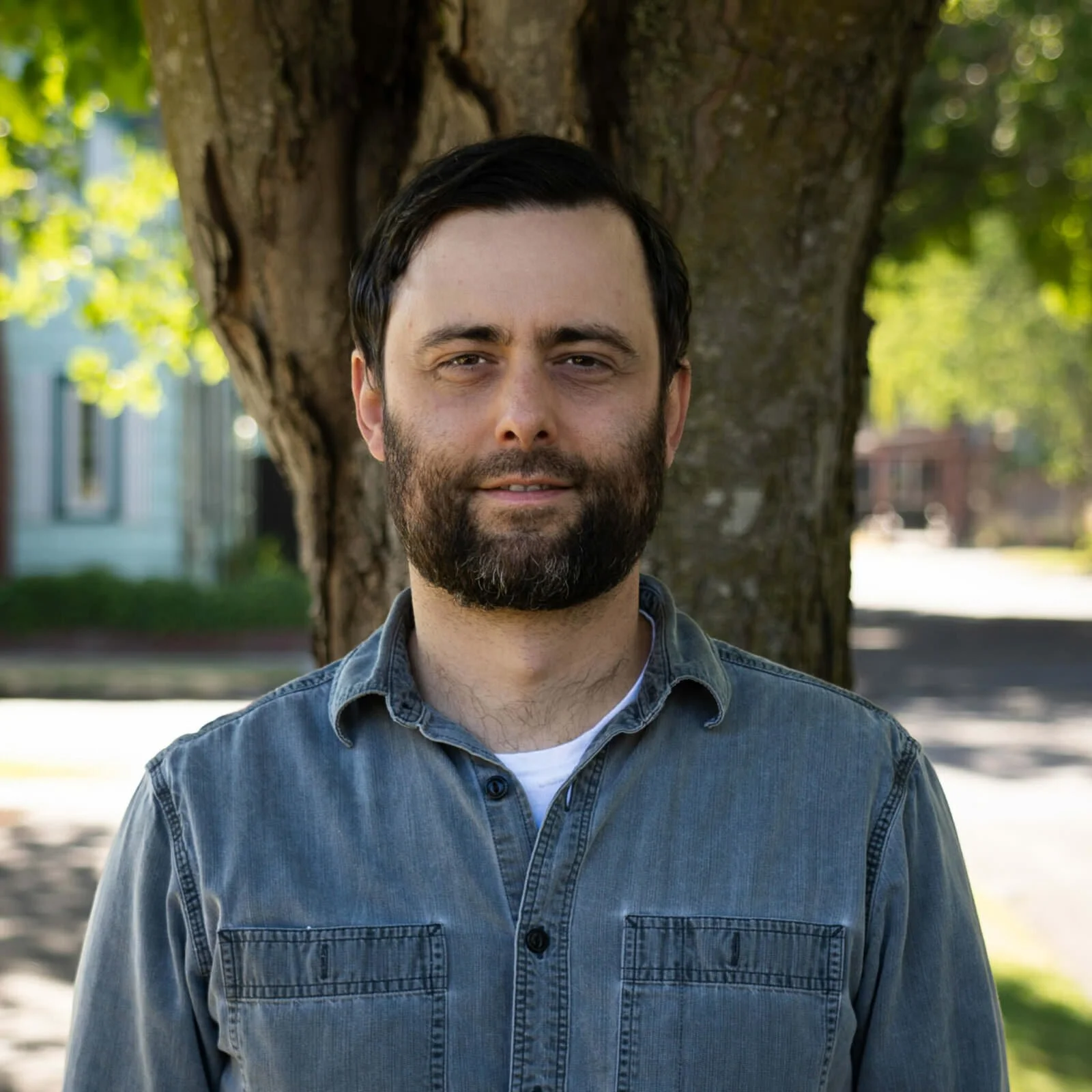 A man with dark hair and a beard standing outdoors in front of a tree, smiling slightly. He is wearing a denim shirt with a white undershirt, and the background includes green foliage and a sidewalk.
