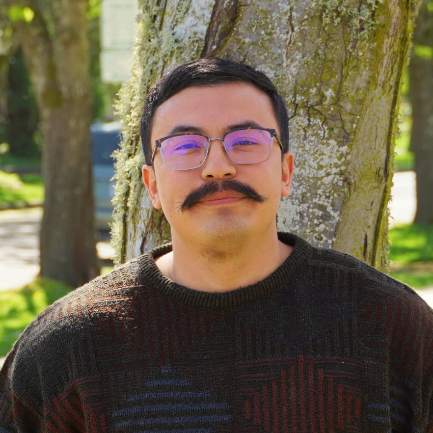Man with glasses, mustache, and light blue shirt standing outdoors near a large tree with green leaves in the background.
