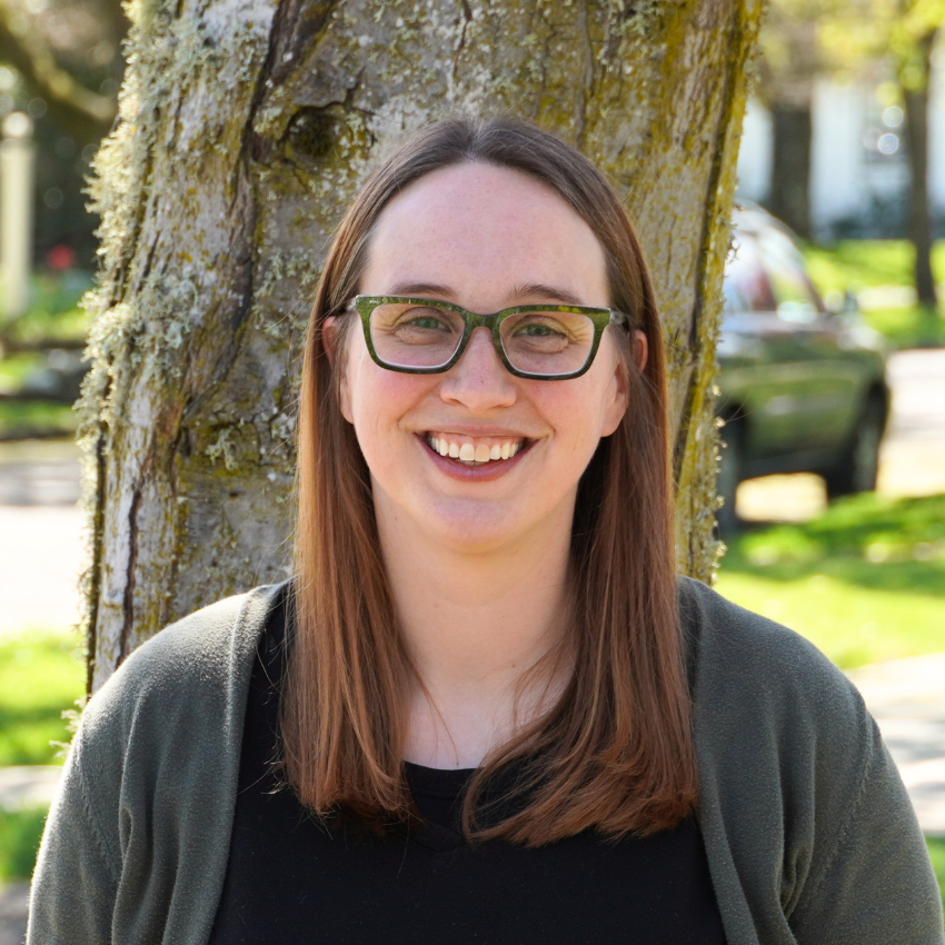 A smiling woman wearing glasses stands in front of a tree outdoors.