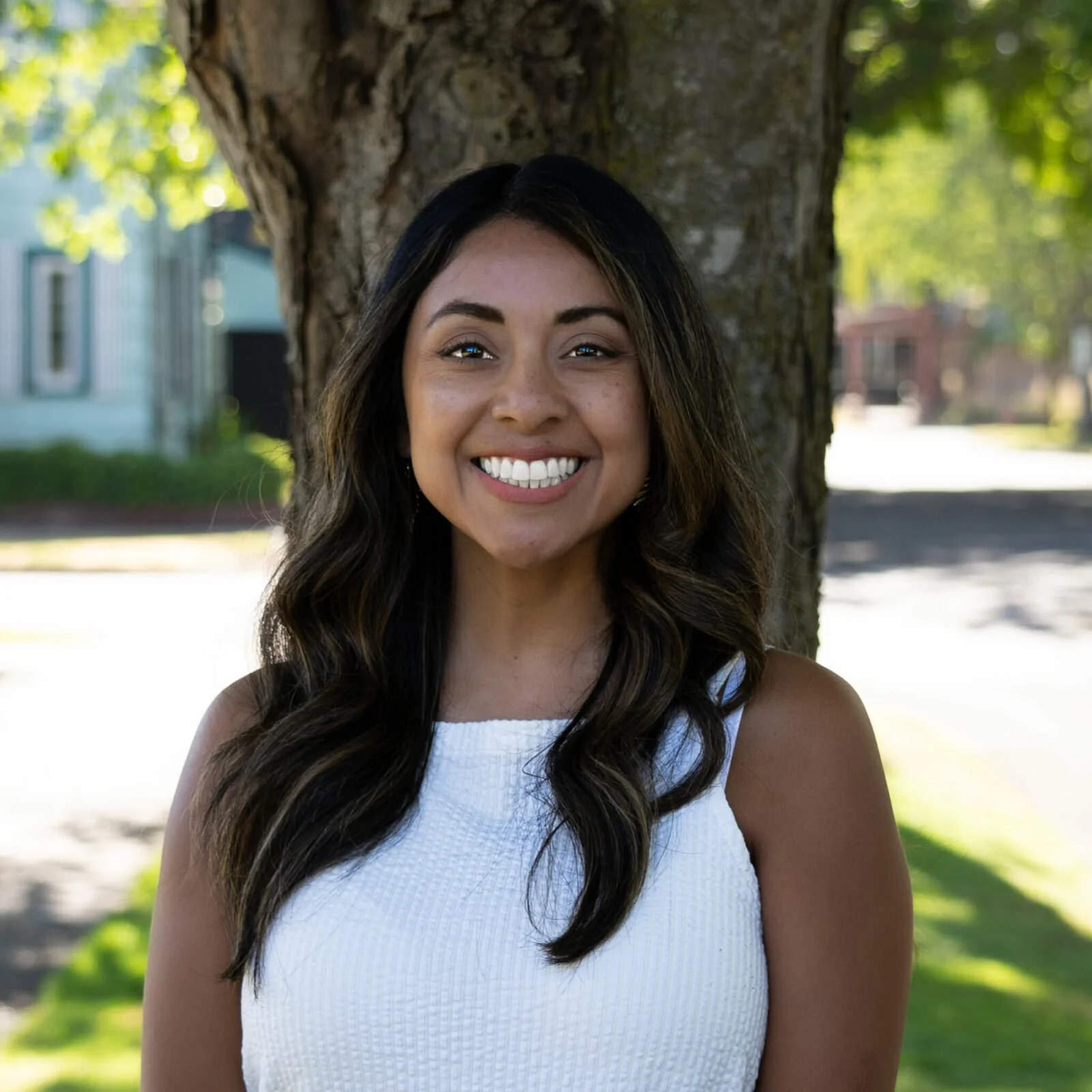 A woman smiling outdoors in front of a tree, wearing a white sleeveless top.