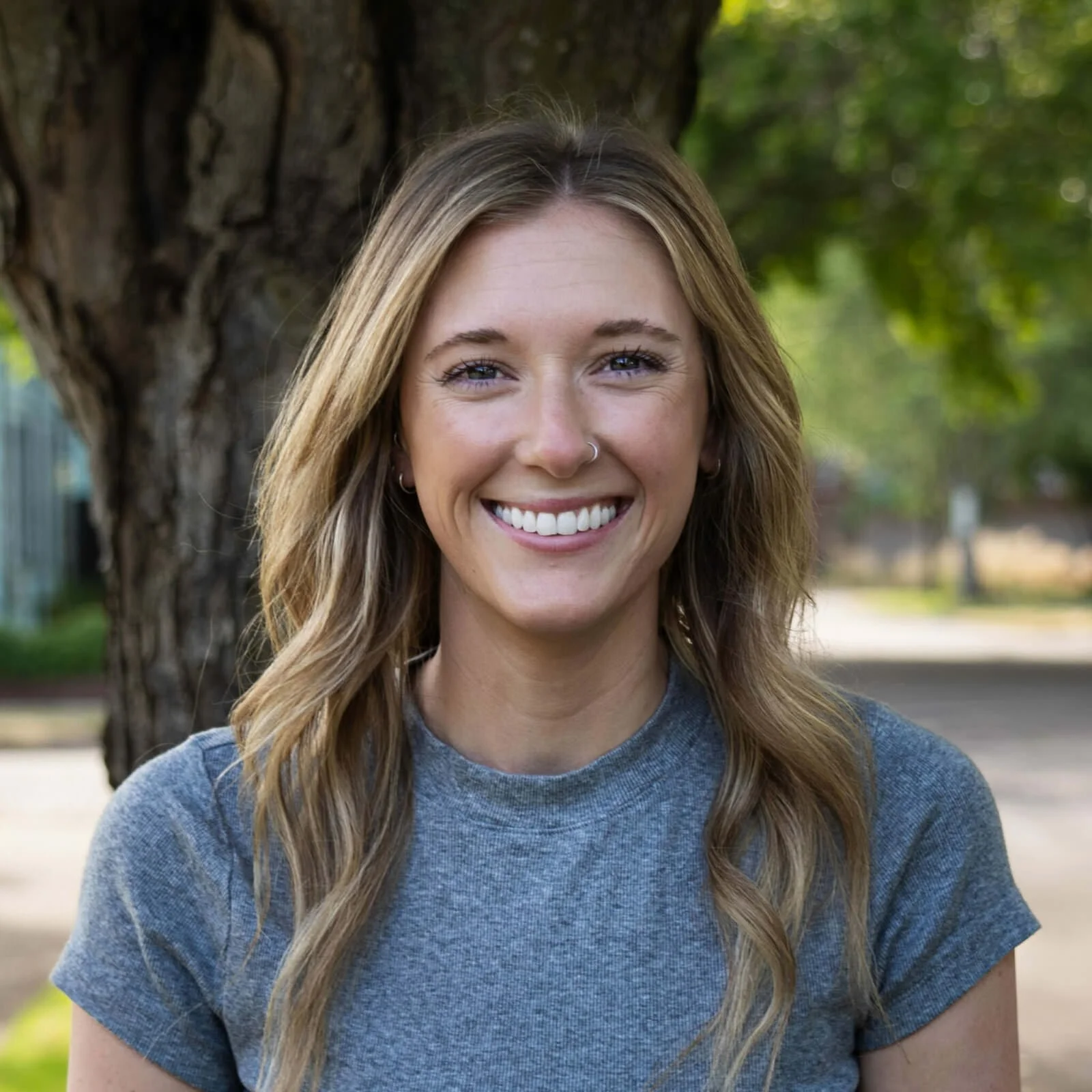 A young woman with long, wavy blonde hair and a nose piercing smiling outdoors in front of a large tree and green foliage.