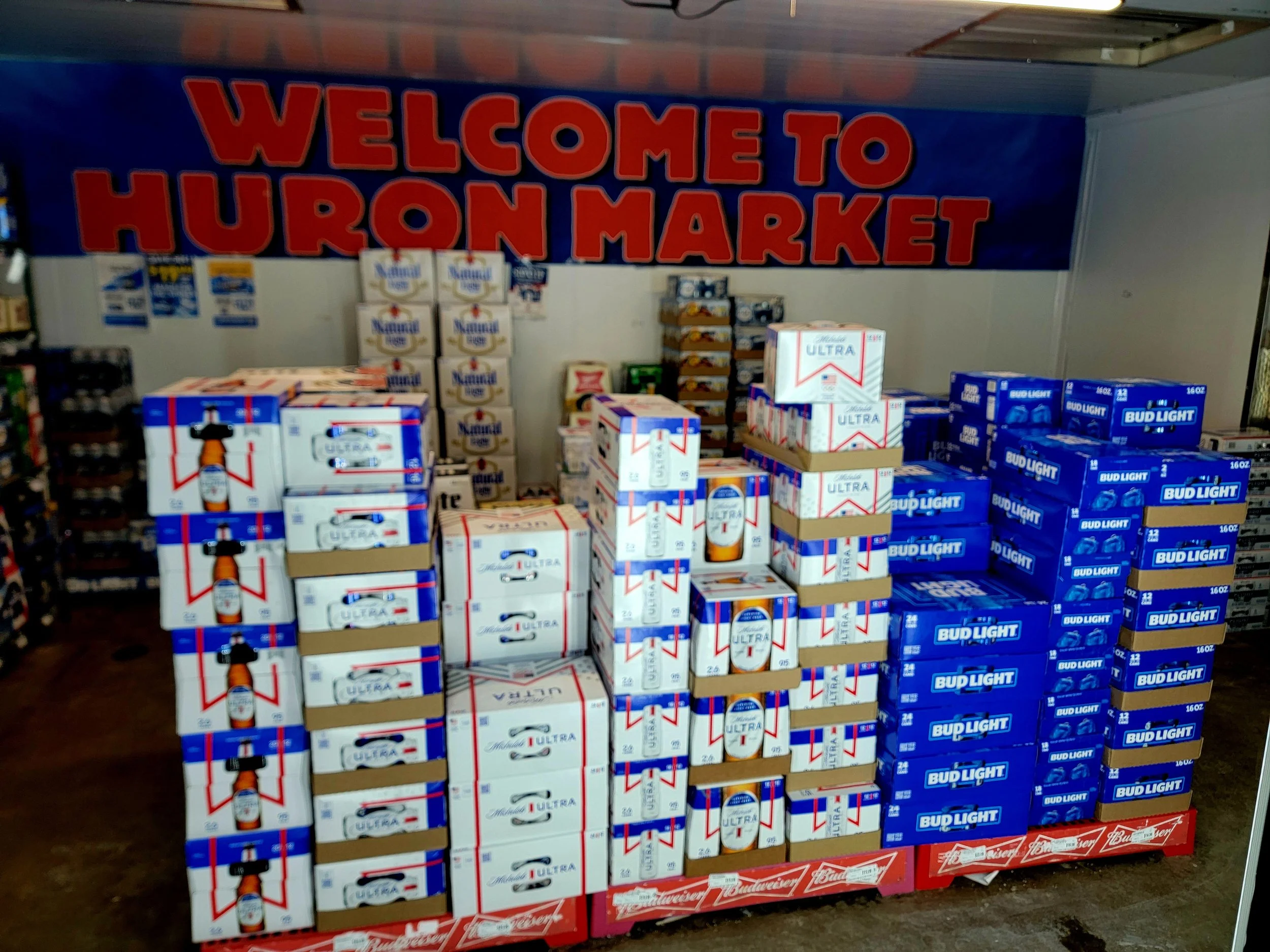 Display of Bud Light beer cases for sale in a store with a large banner above that reads 'Welcome to Huron Market'.