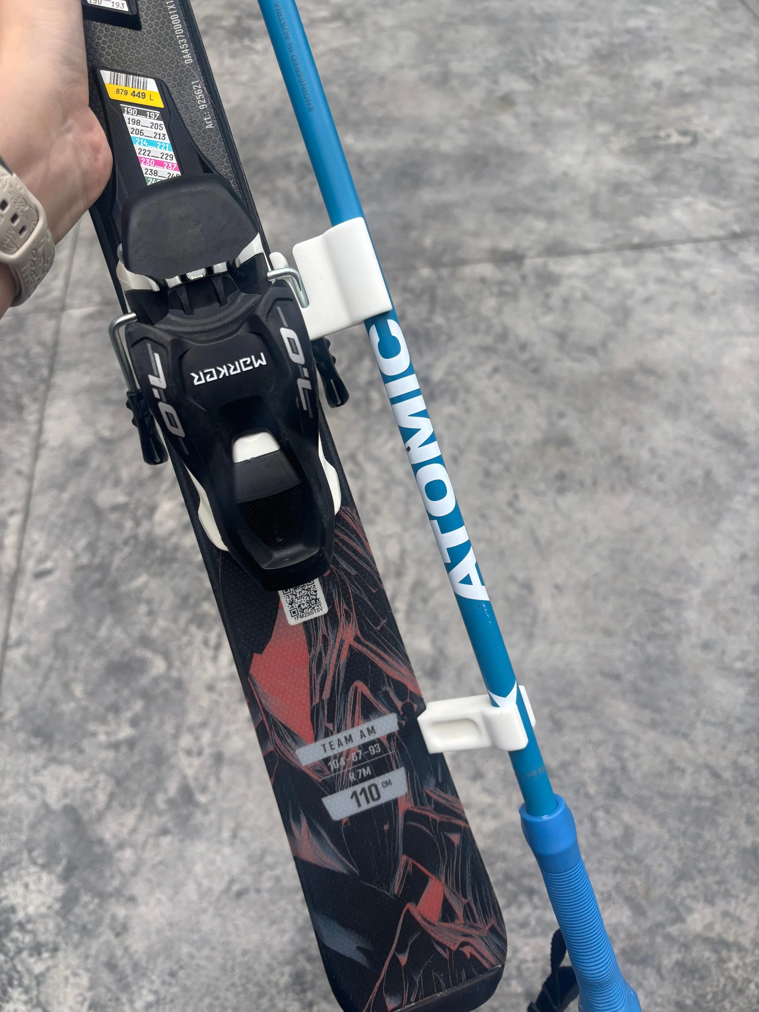 Close-up of a ski with attached ski bindings and a ski pole, held over a concrete surface.