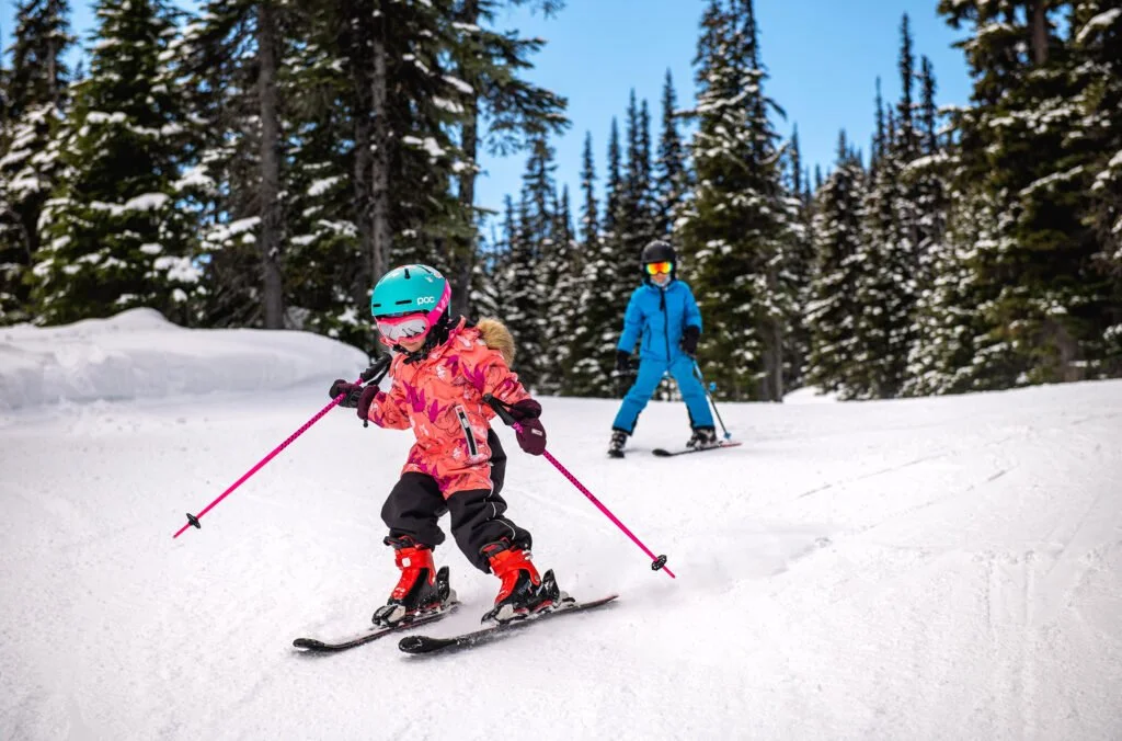 A young girl in a pink jacket and helmet skis down a snowy slope with an adult skier in a blue jacket and pants nearby, surrounded by snow-covered pine trees.