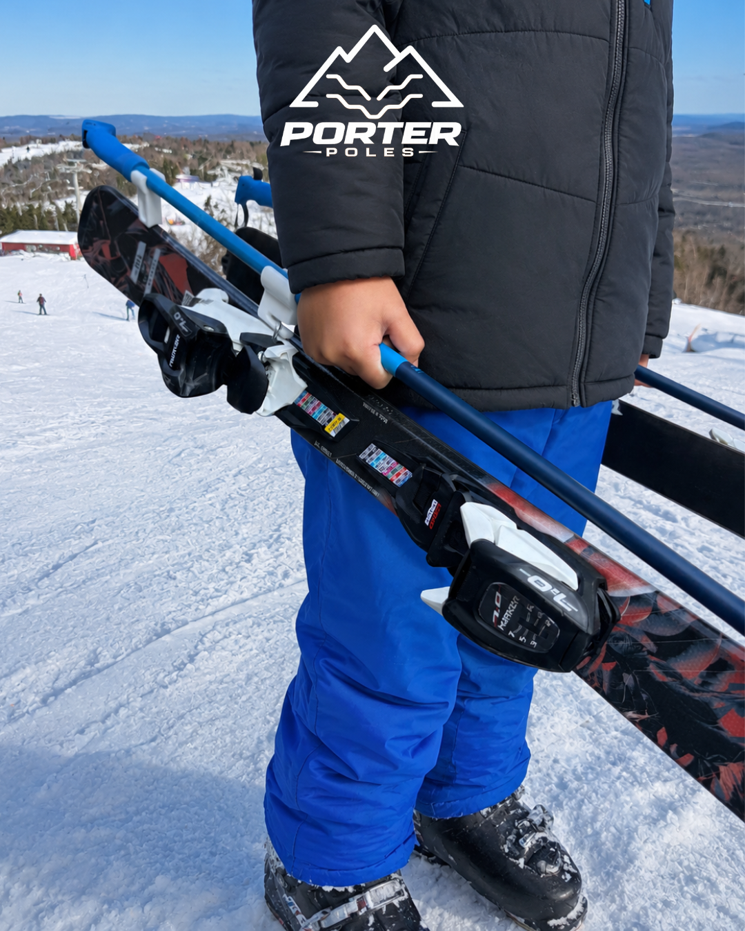 Child holding ski poles at a snowy ski resort with other skiers in the background and a mountain landscape.