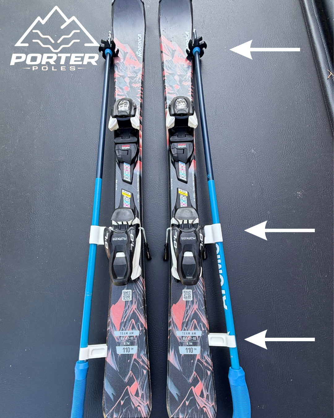 A pair of cross-country skis with poles laid side by side on a black surface. The skis are black with red and gray graphics, equipped with bindings for ski boots. The poles are blue with black handles and straps, attached to the skis with white clips.