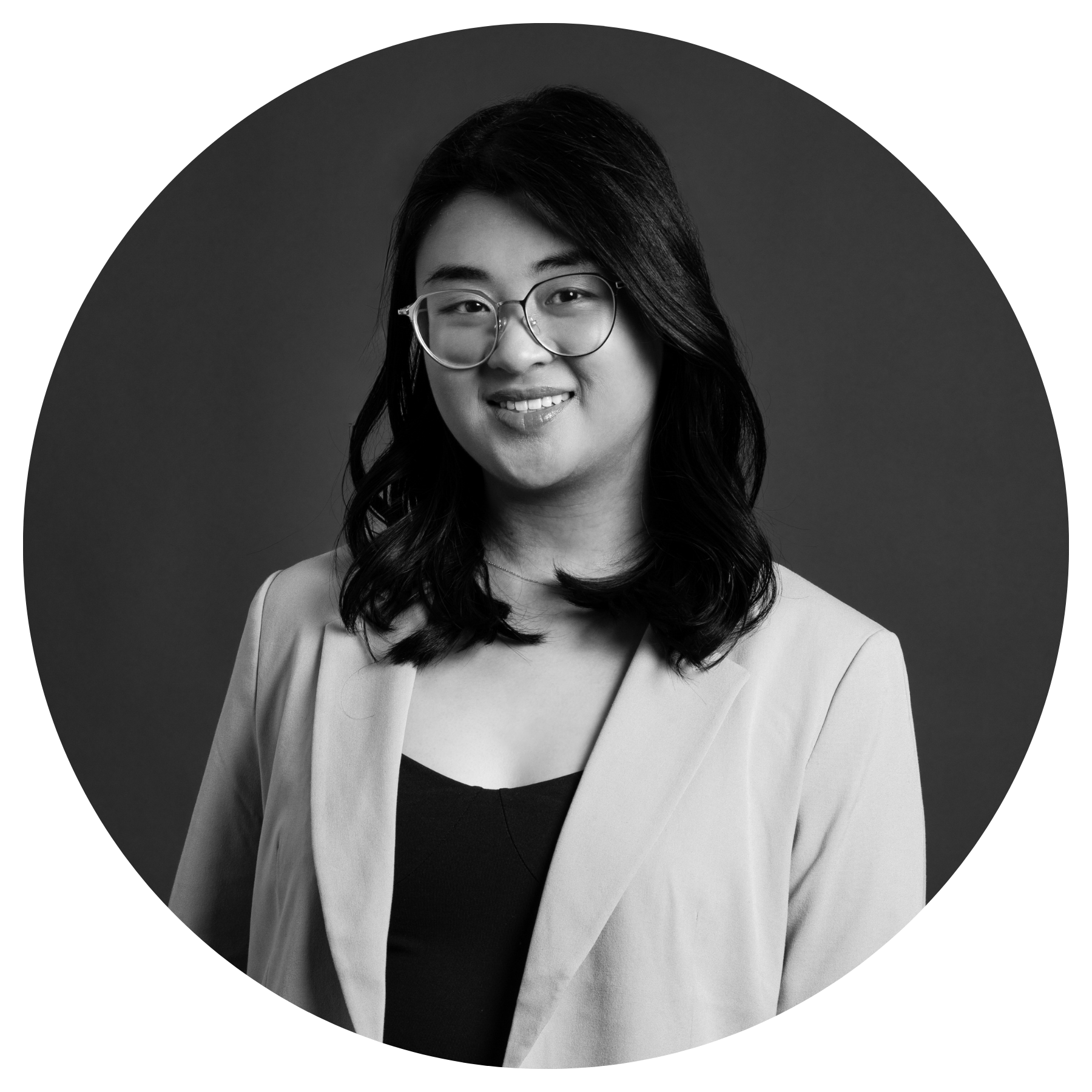Black and white portrait of a smiling woman wearing glasses, a black top, and a light-colored blazer, with wavy hair and a confident expression.