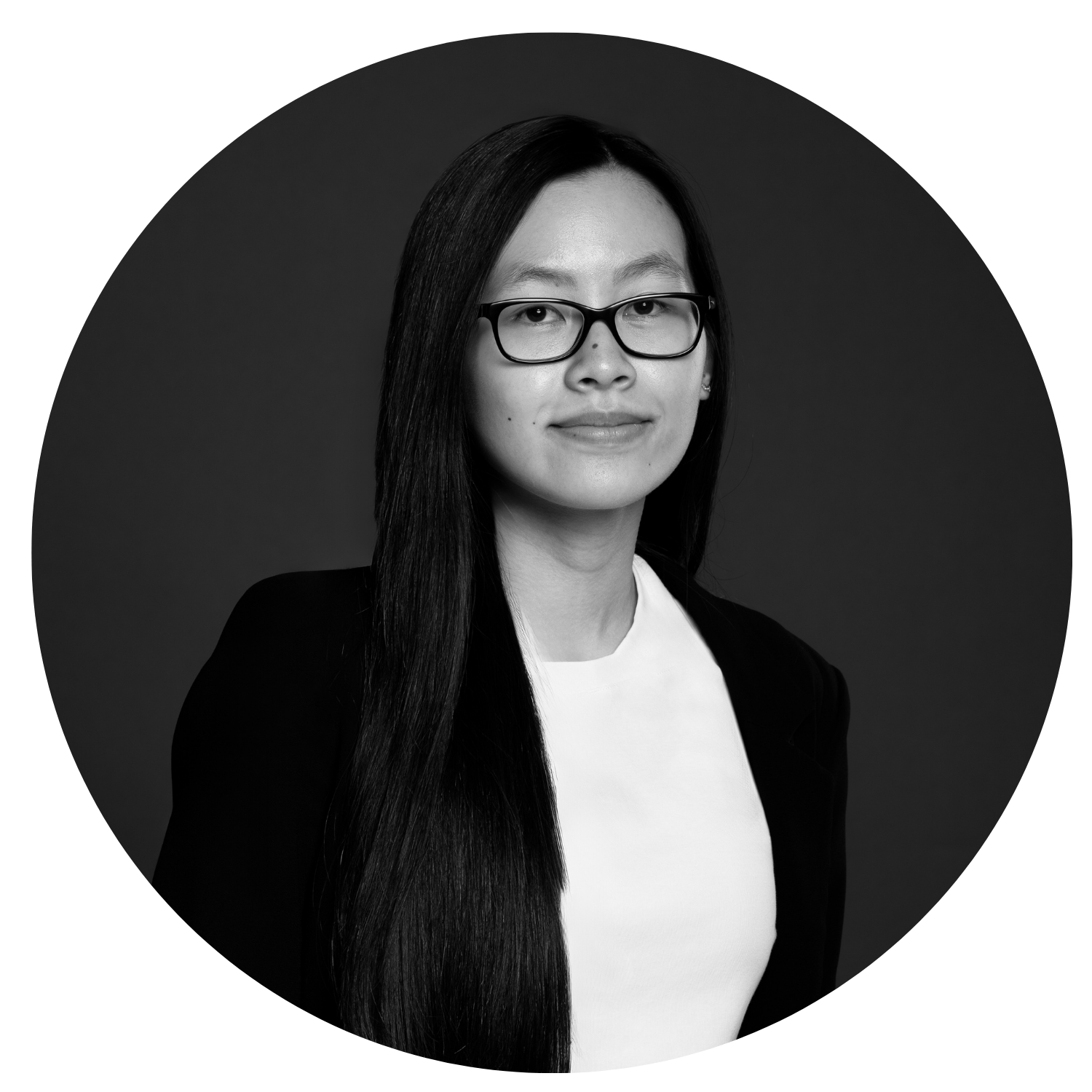 Black and white portrait of a woman with long dark hair, glasses, wearing a white top and dark blazer, looking directly at camera with a slight smile, plain dark background.