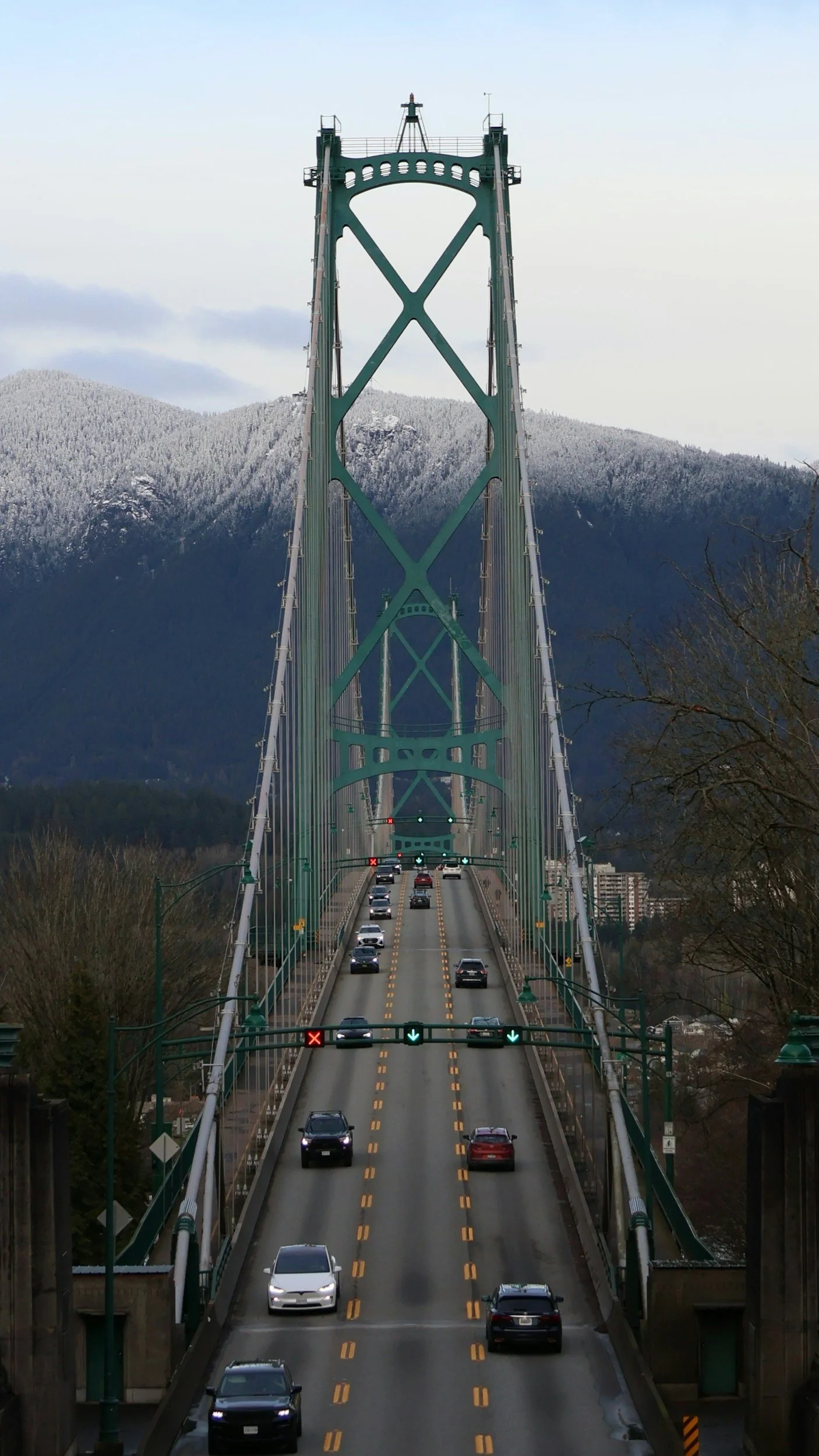 A view of a large green suspension bridge with multiple lanes, vehicles, and traffic signals, set against a backdrop of snow-capped mountains and overcast sky.