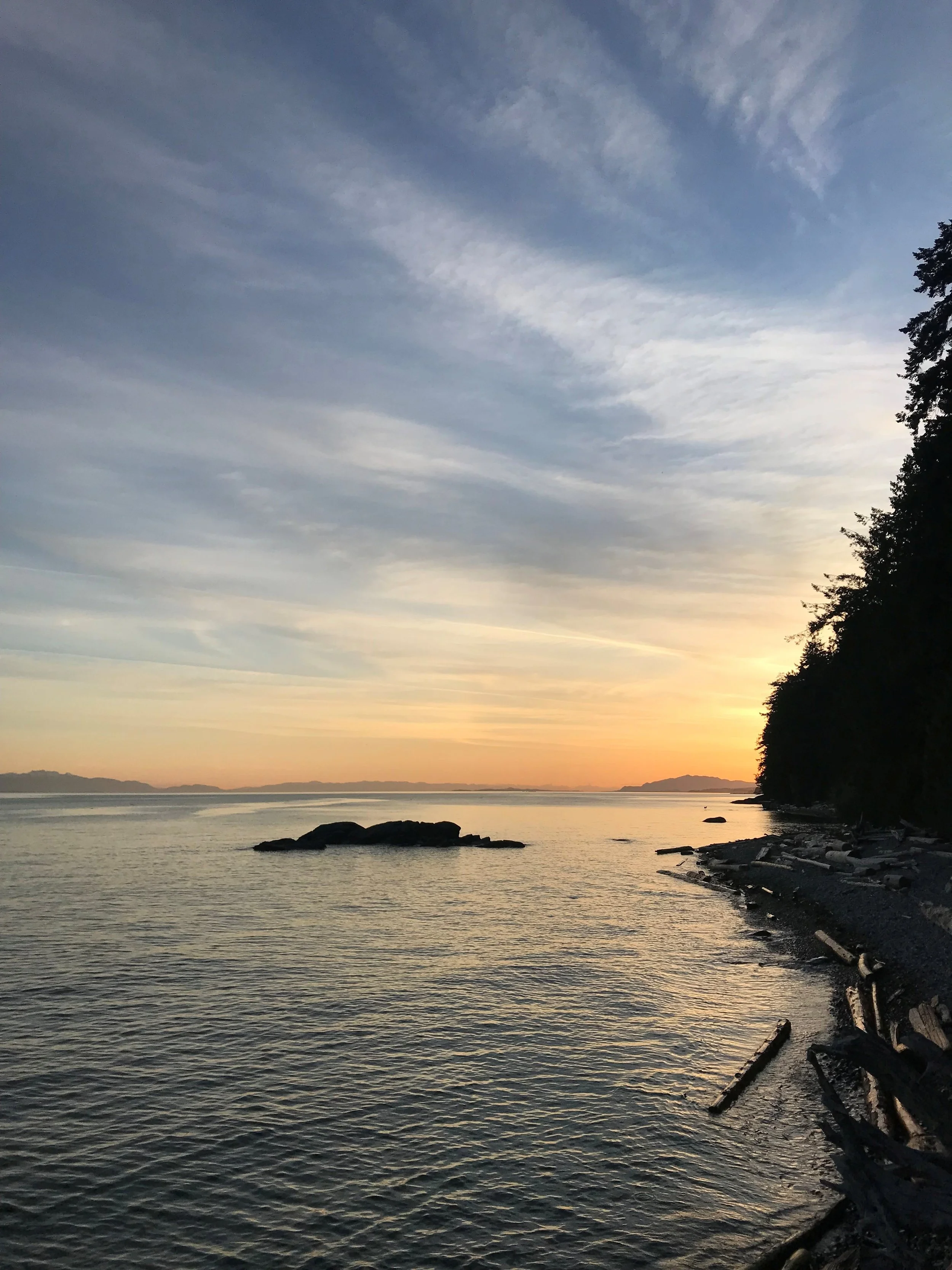 A peaceful sunset over a calm body of water with rocky shoreline and silhouettes of trees on the right.