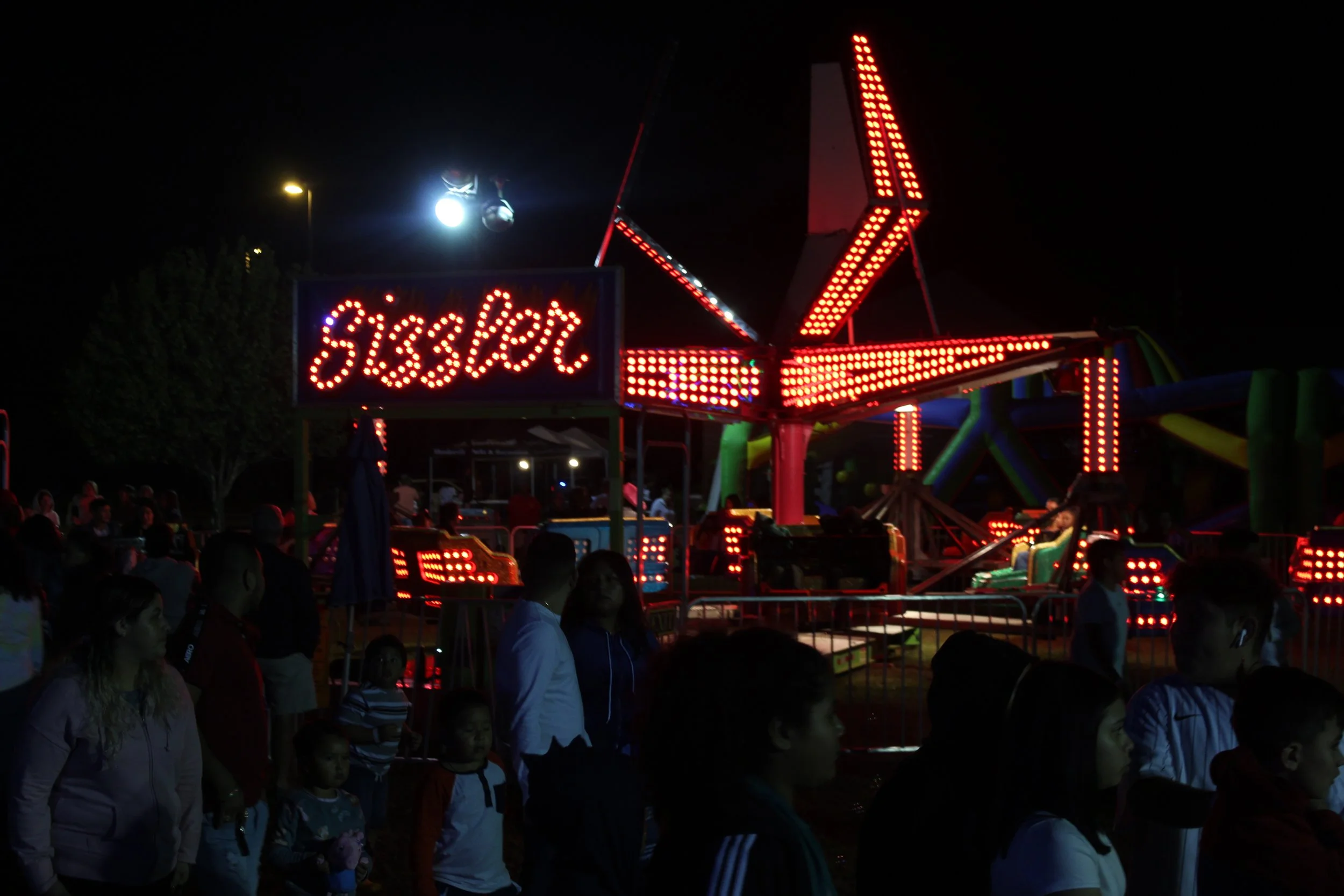 Nighttime scene at a carnival with a crowd of people in front of a brightly lit amusement ride, including a sign that says 'Sizzler'.