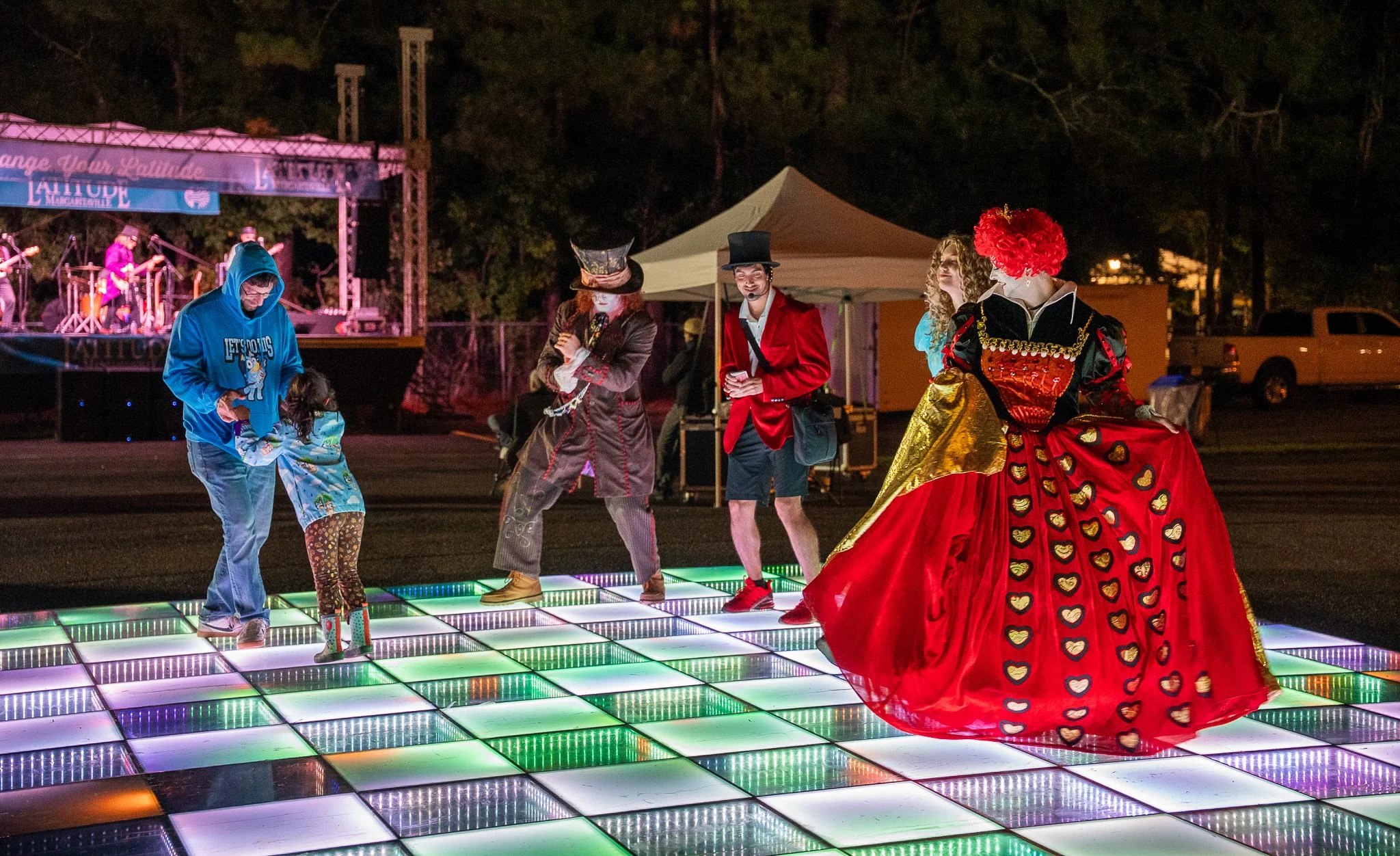 People dressed in costumes dancing on a colorful illuminated dance floor at night, with a stage and band in the background.