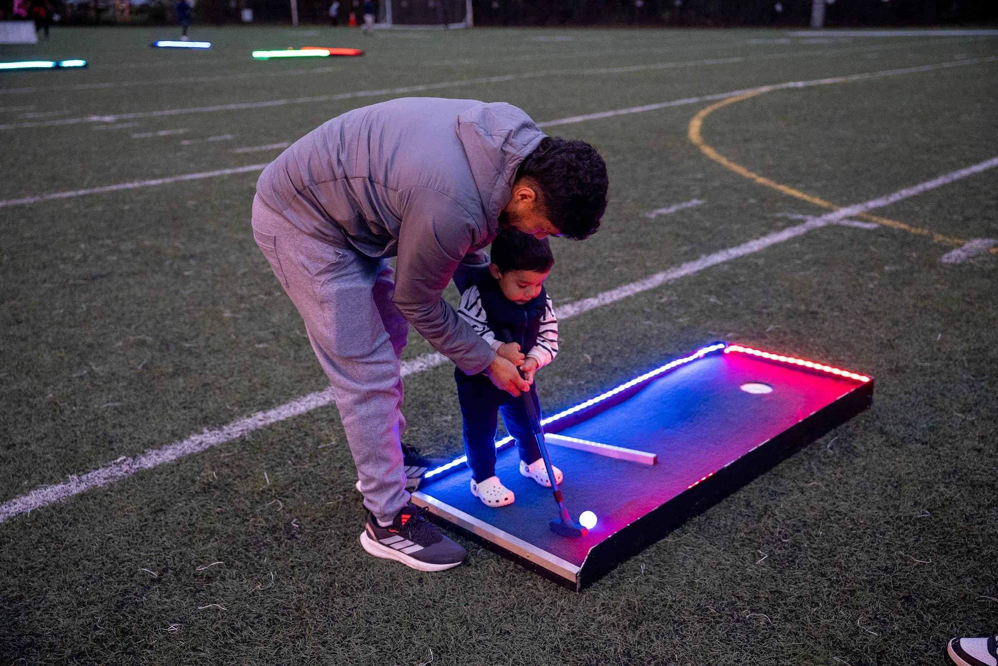 A man and a young boy playing golf on a putting mat outdoors at dusk, with colorful LED lights around the mat and a football field in the background.