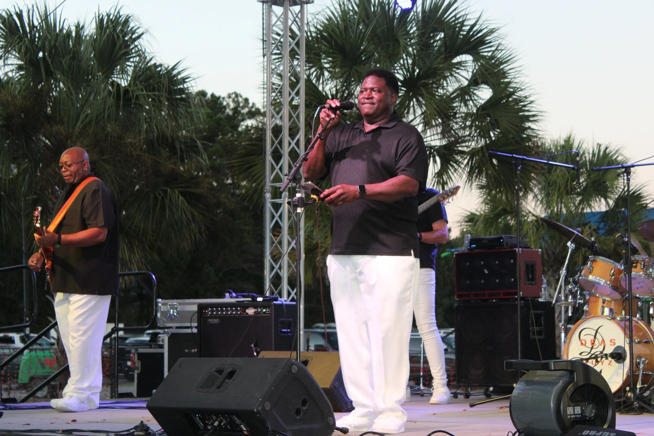 A man wearing a black shirt and white pants is singing into a microphone on an outdoor stage. There are trees with palm-like leaves in the background, and musical equipment such as speakers, guitars, and drums are visible.