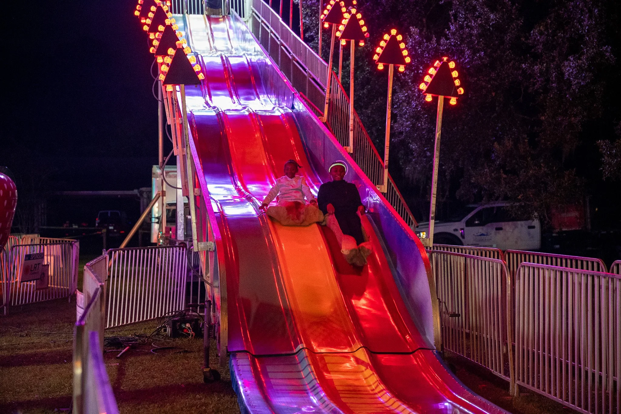 Nighttime scene of a brightly lit amusement park slide with two children sliding down, surrounded by colorful illuminated decorations, and dark trees in the background.