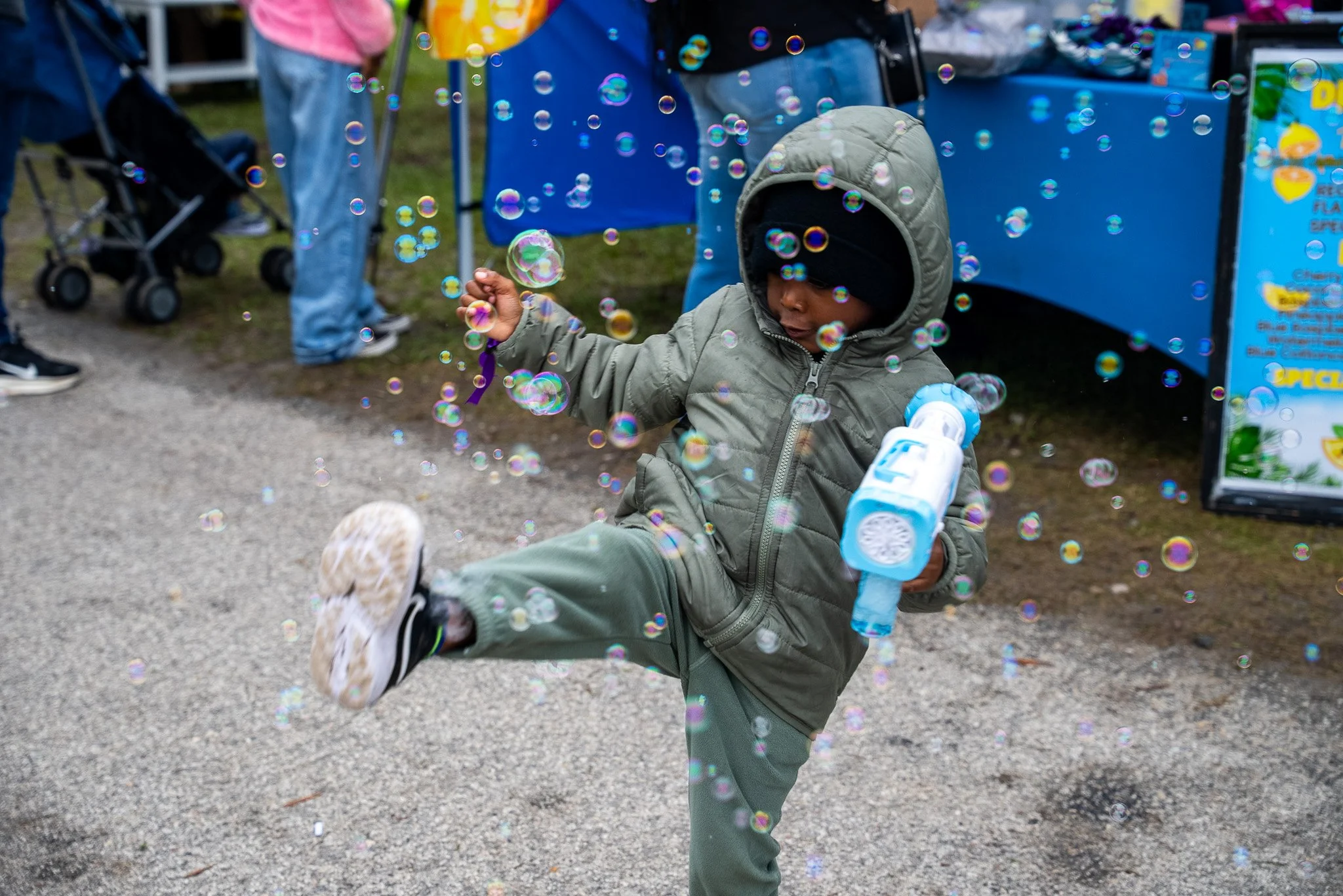 A young child wearing a gray hooded jacket and camouflage shoes plays with bubbles at an outdoor event, with a blue booth and people in the background.