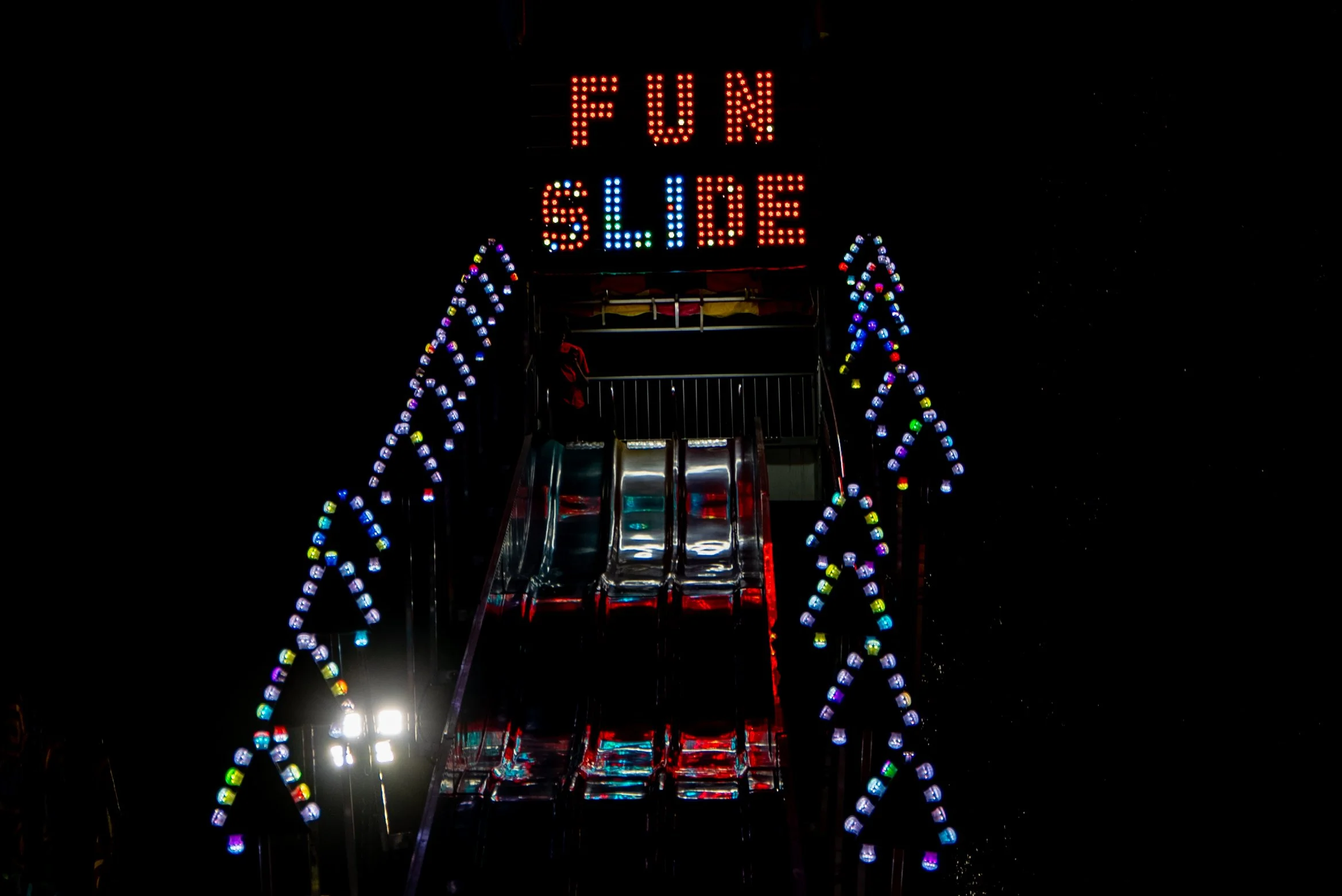 Nighttime view of a carnival ride with illuminated signs reading "FUN SIDE" and colorful decorative lights.