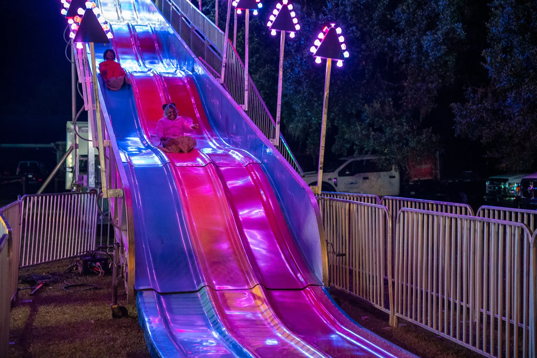 Two children are sliding down a slide that is surrounded by fence barriers, with trees and parked vehicles in the background.