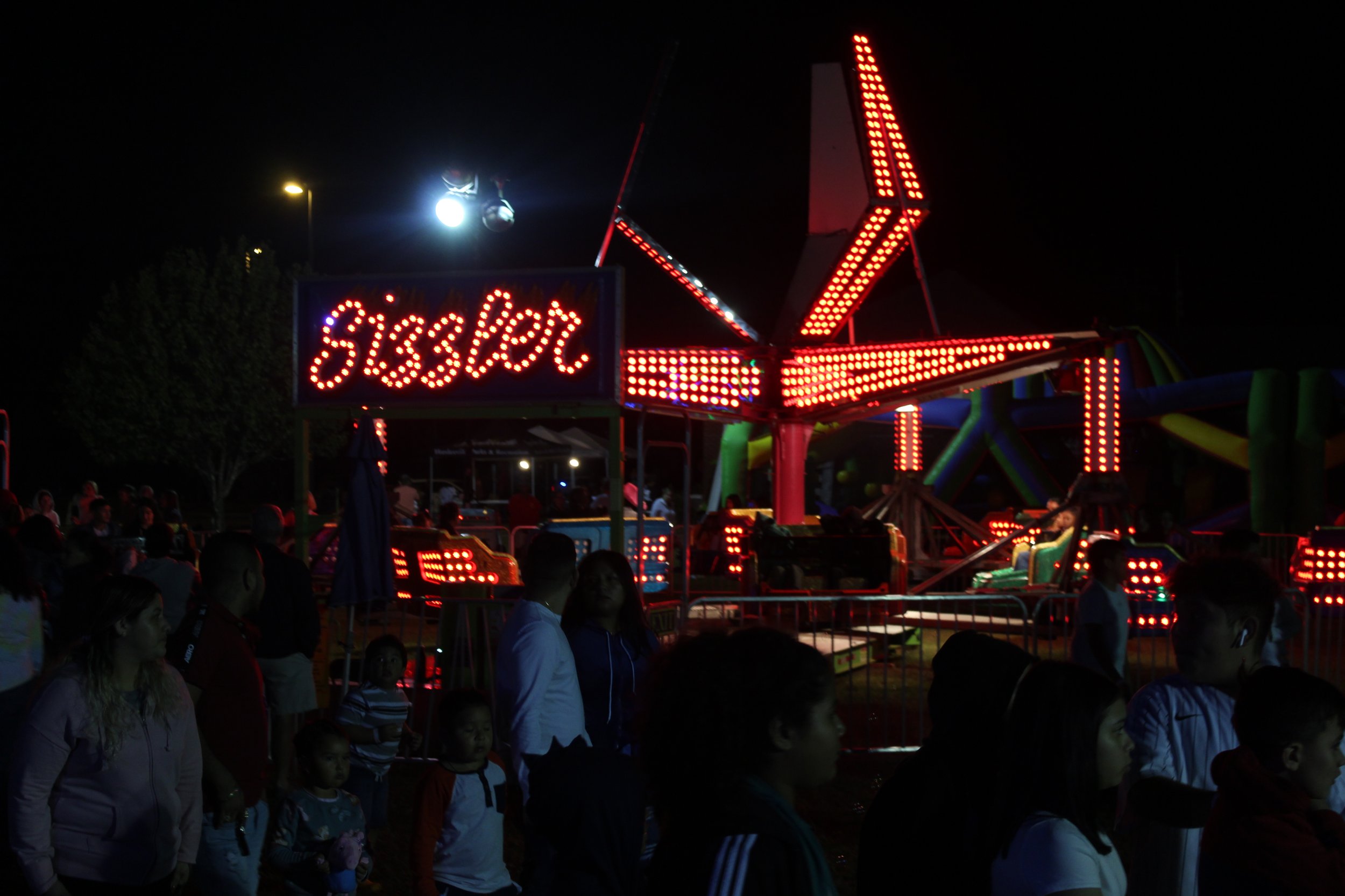 Night scene at a fair with brightly lit rides and people walking around.