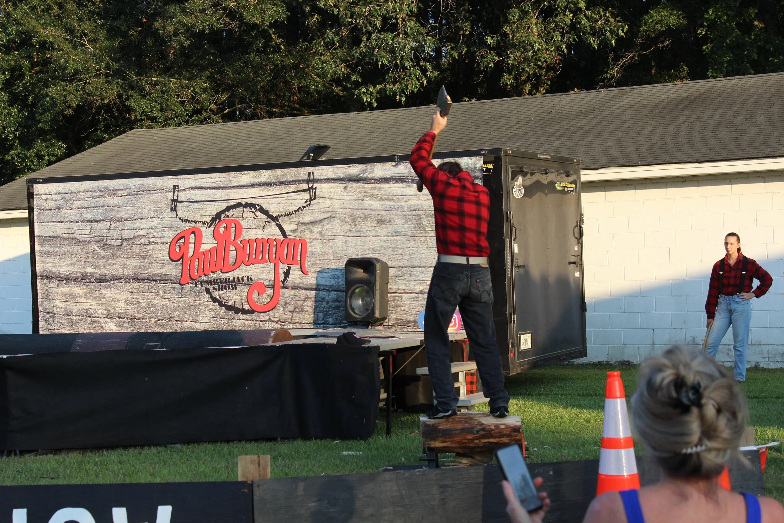 Man in a red flannel shirt throwing a book onto a stage at an outdoor event, with a large sign that reads "Fat Buryan" and a person in a red flannel shirt standing nearby, in front of a building and trees.