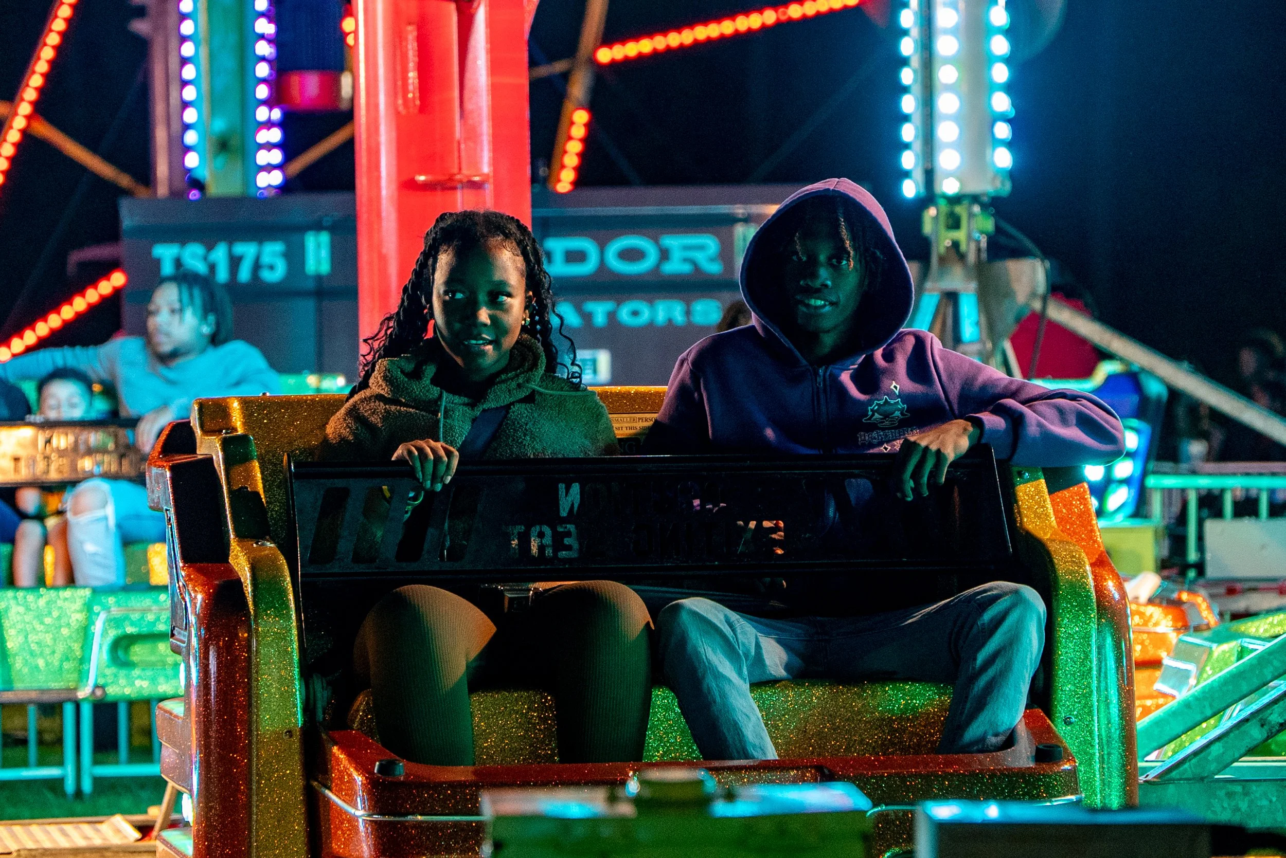 Two children riding a colorful amusement park ride at night, surrounded by bright, flashing lights.