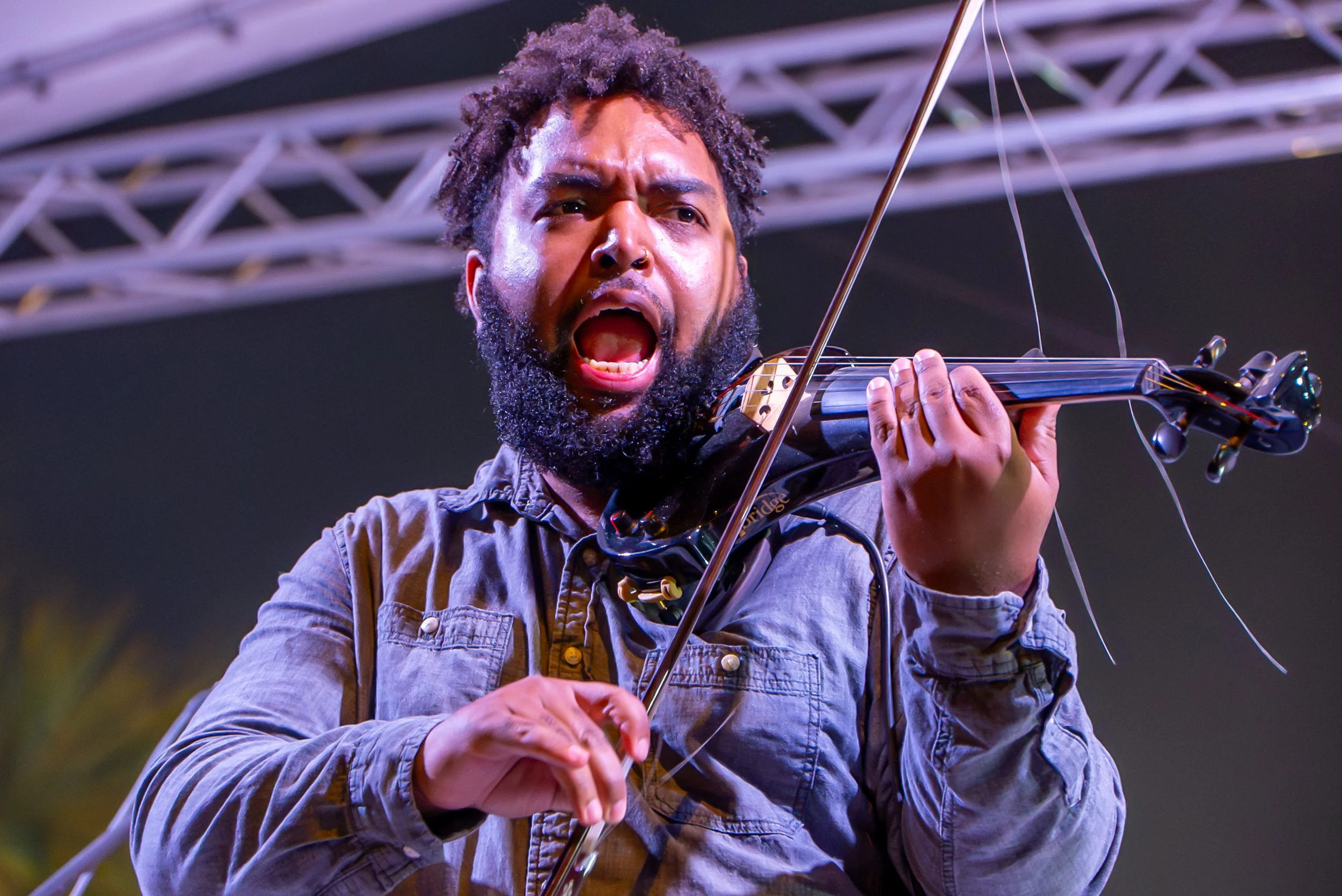 A man with a beard and curly hair passionately playing the violin on stage, with a metal framework in the background.
