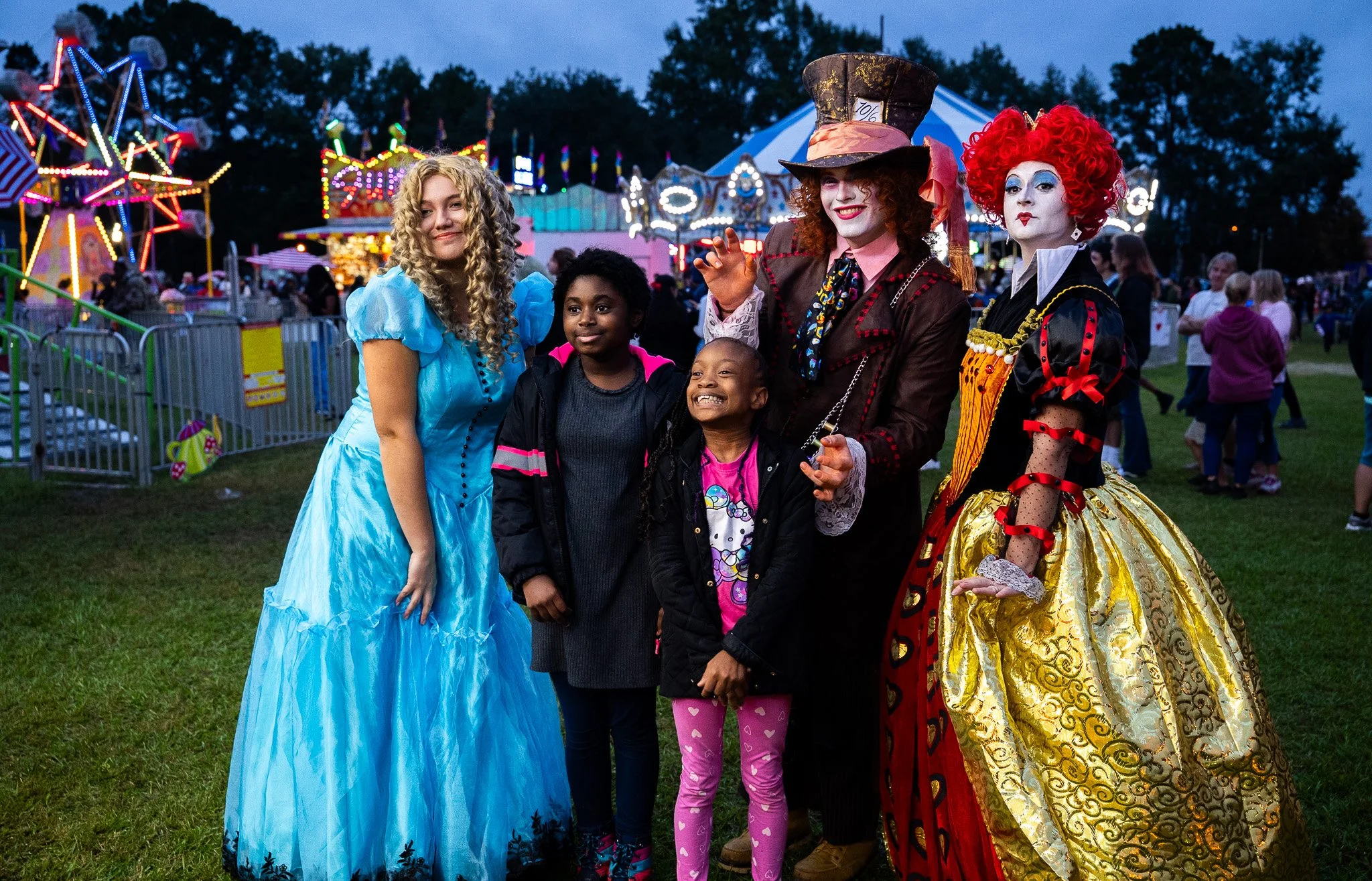 Festival goers dressed as characters from Alice in Wonderland, standing together at a fairground with amusement rides and colorful lights in the background.