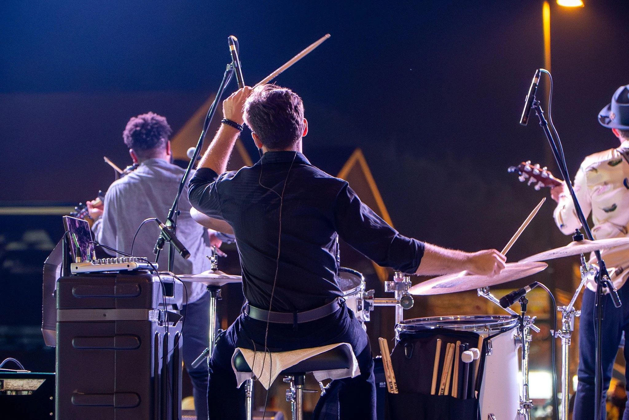 Musicians performing on stage with drums, guitar, and electronic equipment during a concert at night.