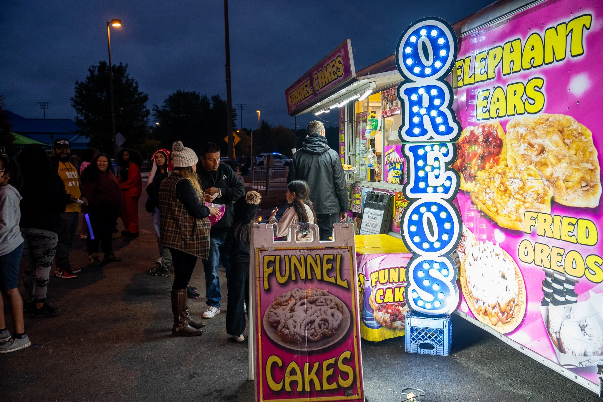 Festival themed food stand at night selling funnel cakes, elephant ears, fried Oreos, and glazed donuts, with customers and colorful signage.