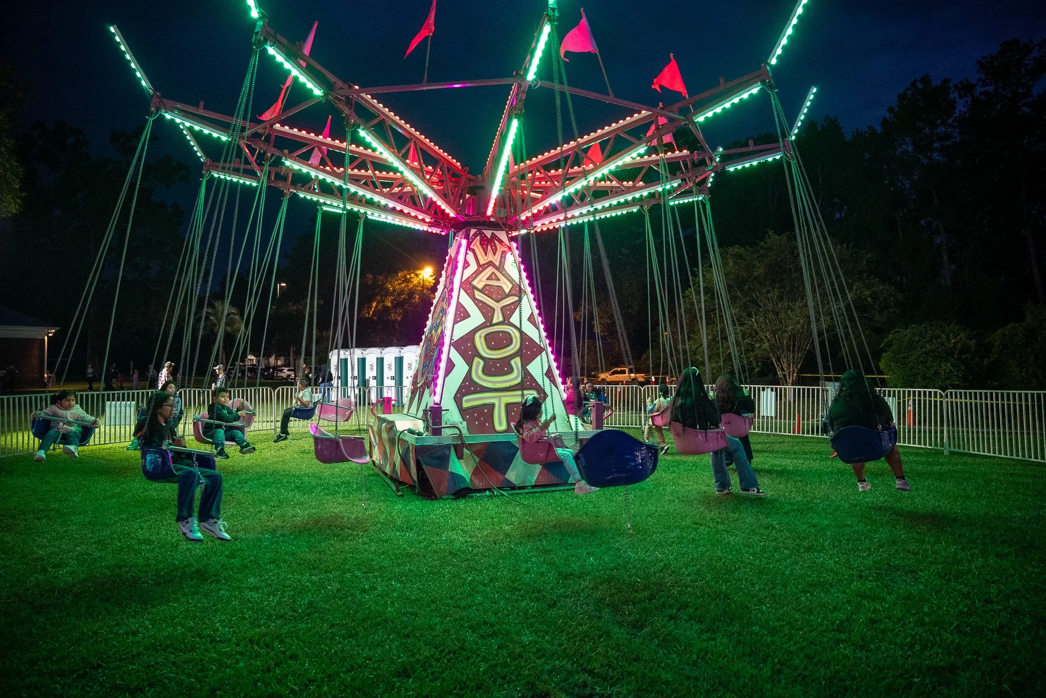 Children riding on a brightly lit carnival swing ride at night, with green and pink lights, surrounded by a fence and trees.