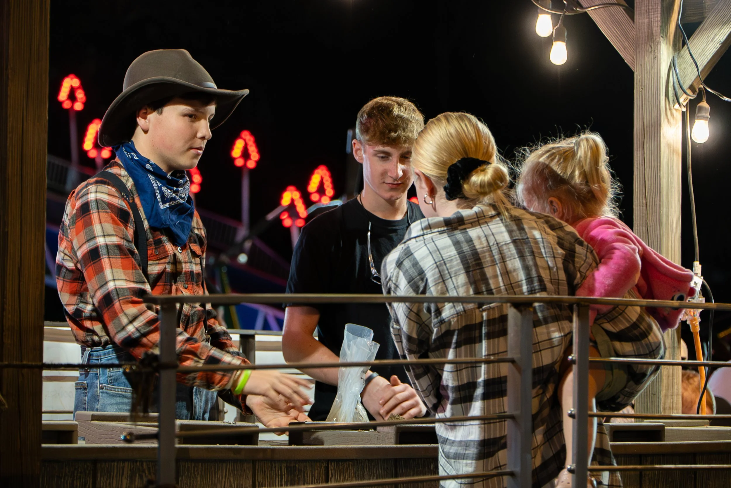 Nighttime scene at a fair or carnival with five people gathered around a game booth, illuminated by hanging light bulbs. The group includes two young women, a young man, and two other individuals, all engaged in a conversation or game, with colorful 