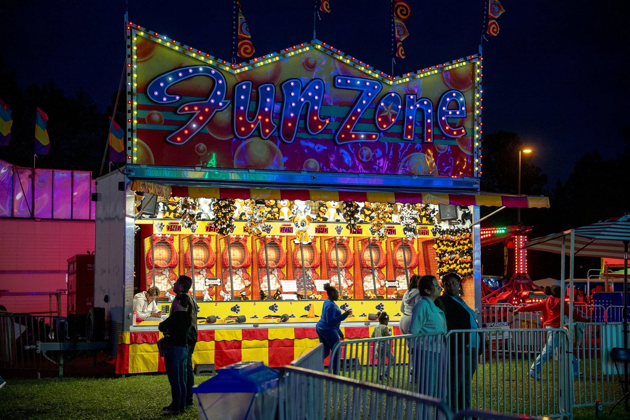 Night scene with a brightly lit sign. Below the sign is a game area with plush toys shaped like dogs and other animals as prizes. Several people are seen walking and standing around the area. A fence surrounds the carnival rides and game booths.