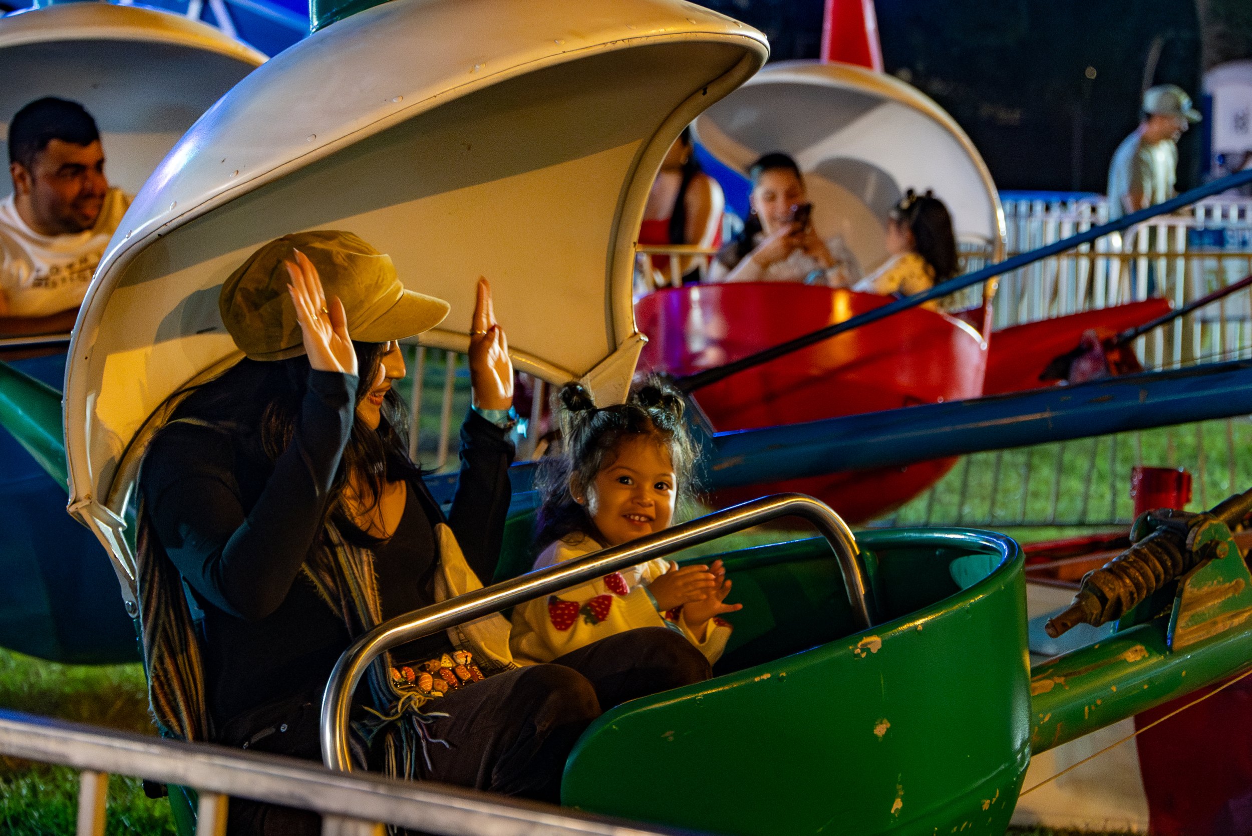 Children riding a colorful amusement park ride at night. The focus is on a girl in a green ride vehicle smiling, with other children and a woman wearing a hat and a child taking a photo in the background.