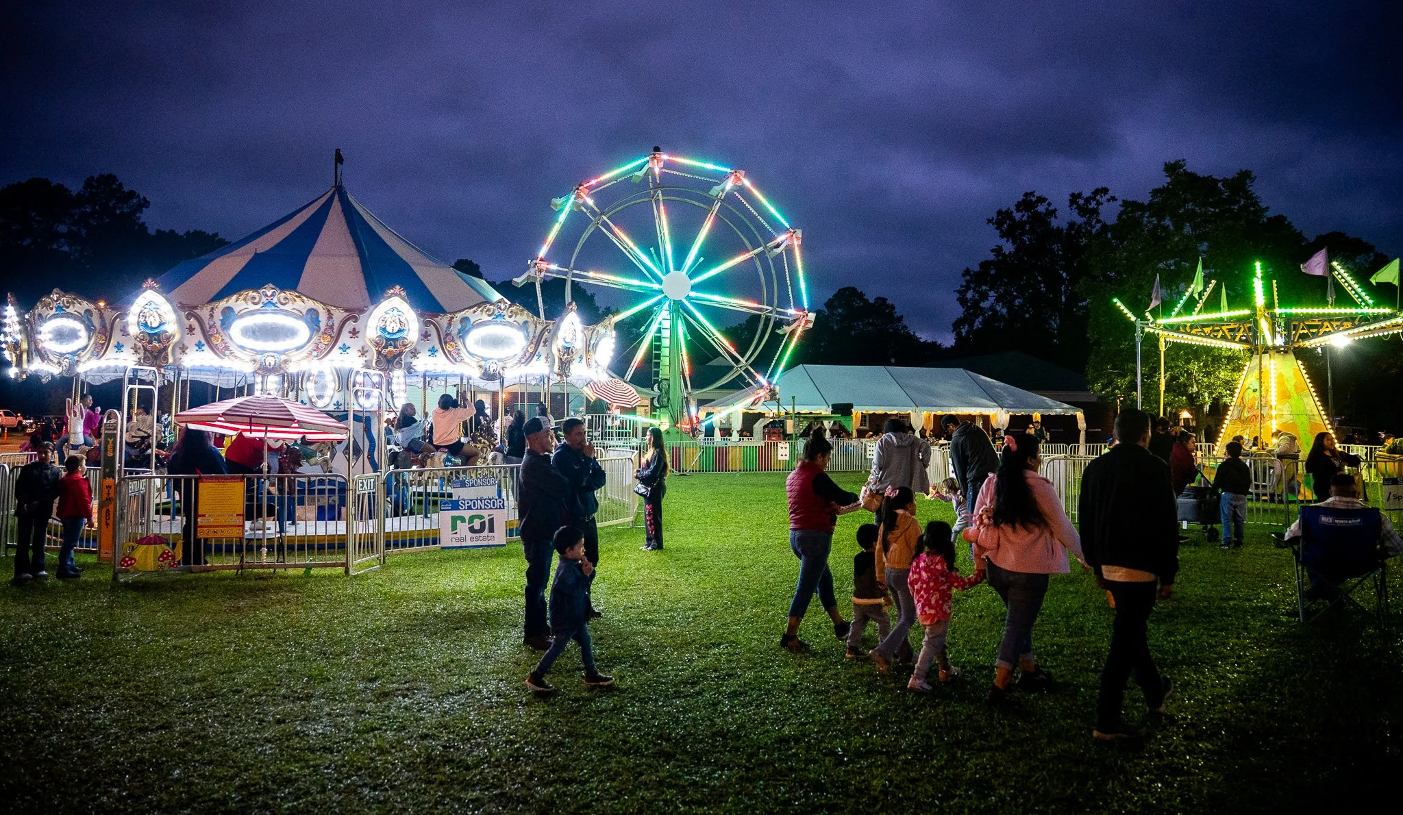 A lively amusement park at night with a Ferris wheel lit up with colorful lights, a carousel, and a small swing ride. Many people, including children and adults, are gathered on the grass field, enjoying the attractions and the festive atmosphere.
