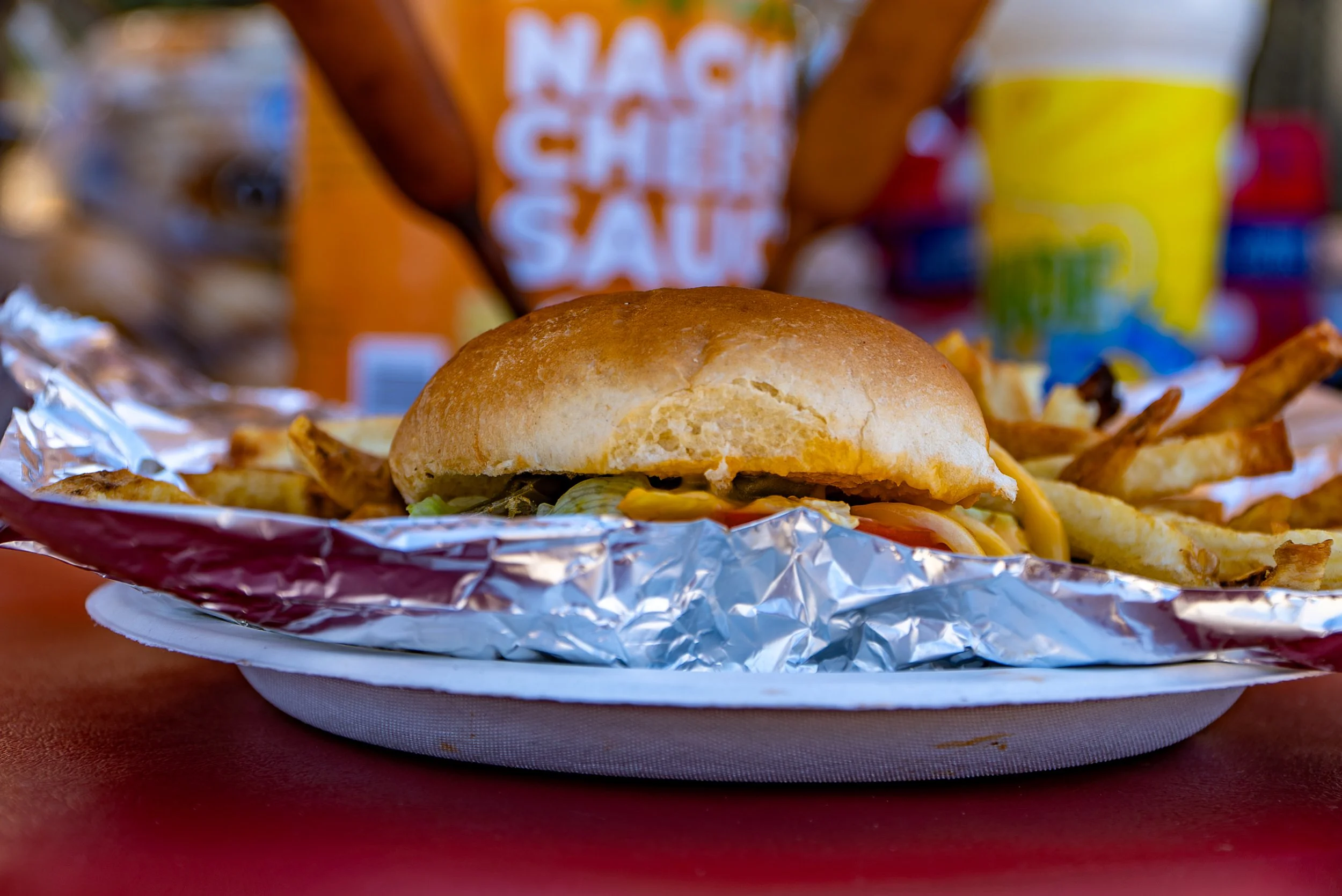 Close-up of a cheeseburger with fries on foil, with a bag of nacho cheese and a yellow cup in the background.