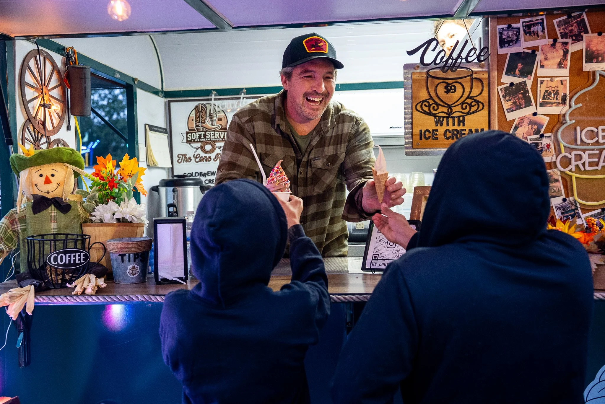 A man serving ice cream at a stall, with children in hoodies in front of him. The stall decor includes a scarecrow decoration and various signs.