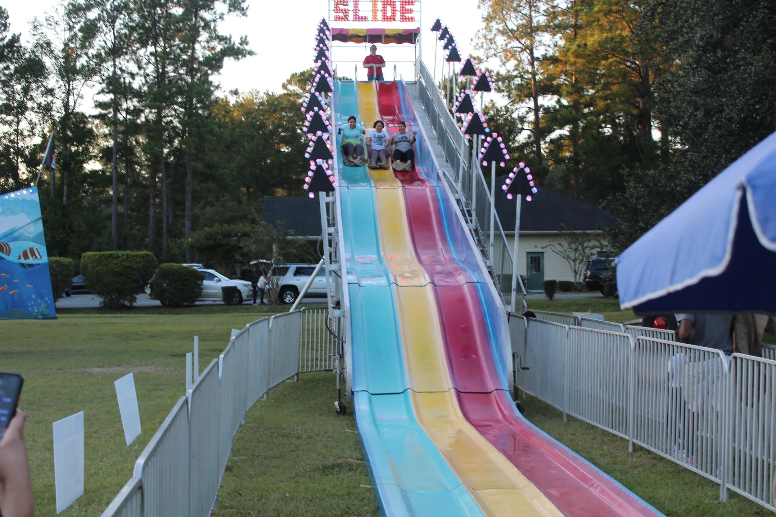 Three people riding on colorful slides at an amusement park, with a person standing at the top of the slide area, surrounded by trees and a parking lot in the background.