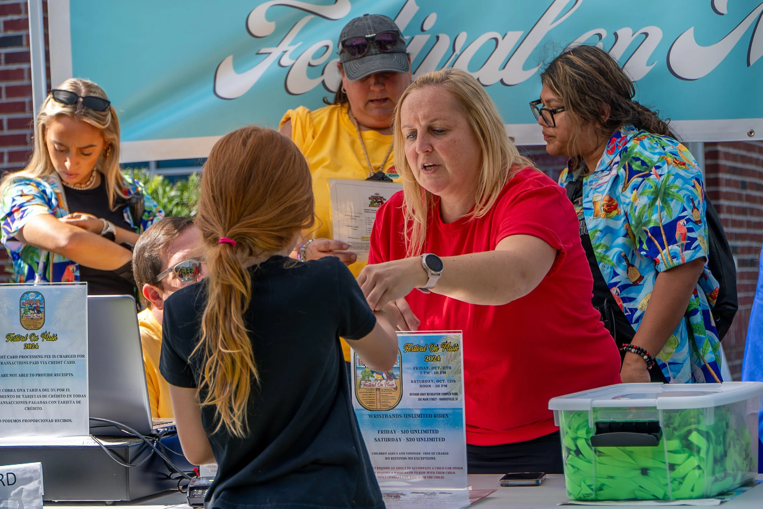 Group of people at a registration table for a festival, with signs and a colorful backdrop, outdoors during daytime.