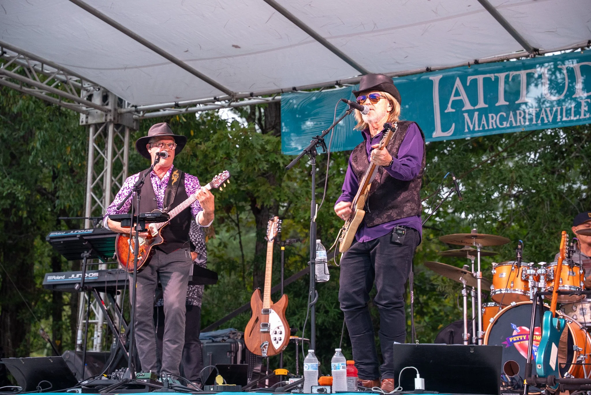 Two male musicians are performing on an outdoor stage, playing guitars and singing into microphones. The stage has a banner that reads 'LATITUDE MARGARITAVILLE' and is set among trees, with musical equipment and water bottles visible in the foregroun