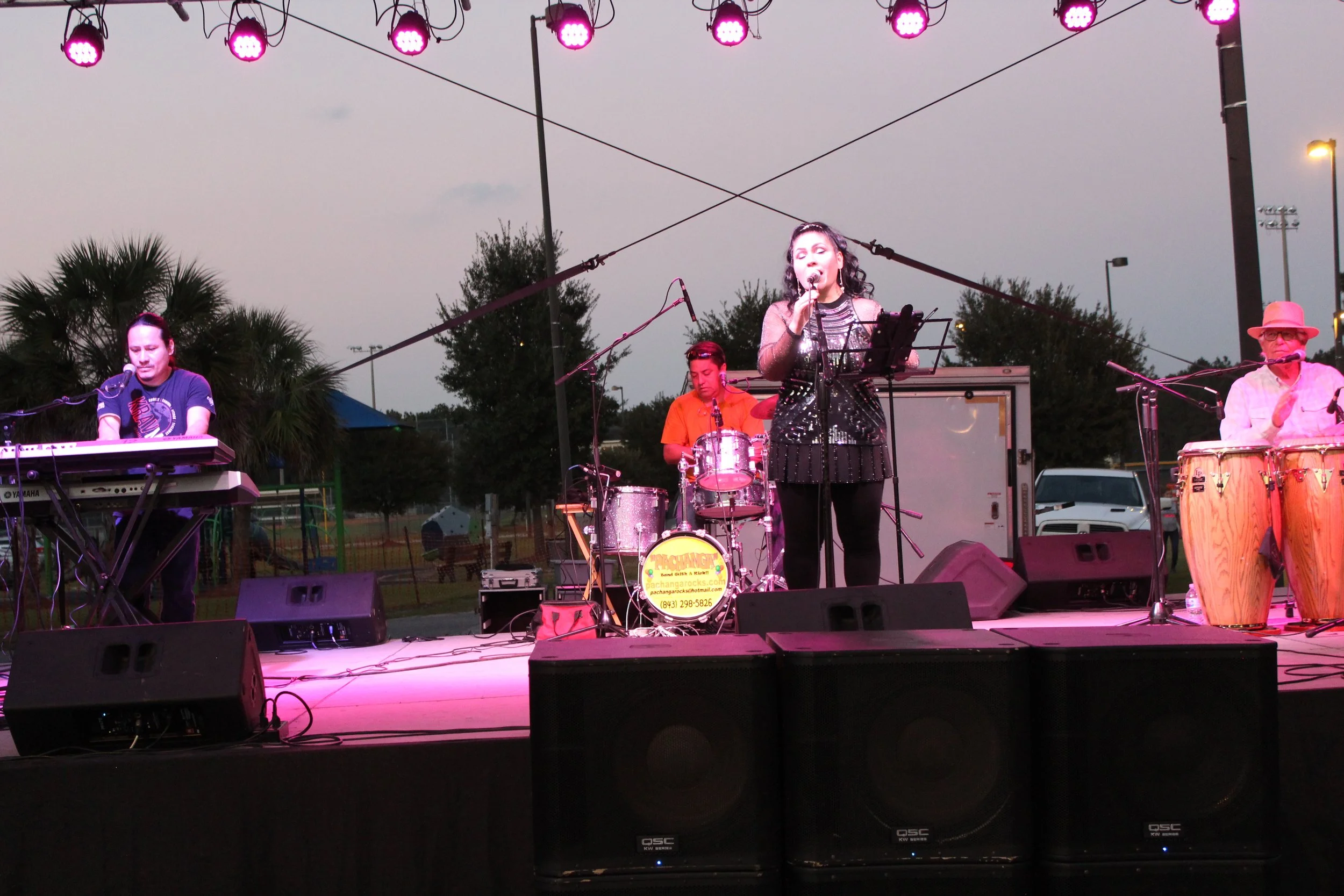 A live outdoor musical performance with four musicians on stage under pink stage lights, including a female singer in the center, a keyboardist on the left, a drummer behind the singer, and a percussionist on the right wearing sunglasses and a hat.