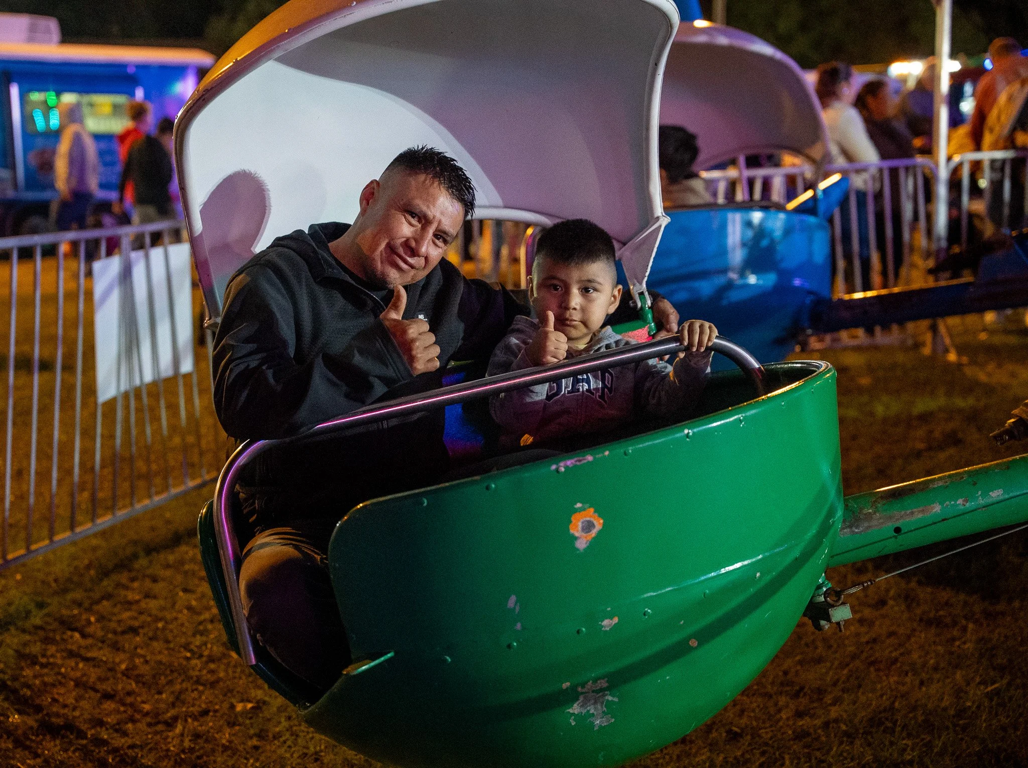 A man and a young boy giving thumbs-up inside a green amusement park ride car at night. The ride is surrounded by a white metal fence with other park attractions and groups of people in the background.
