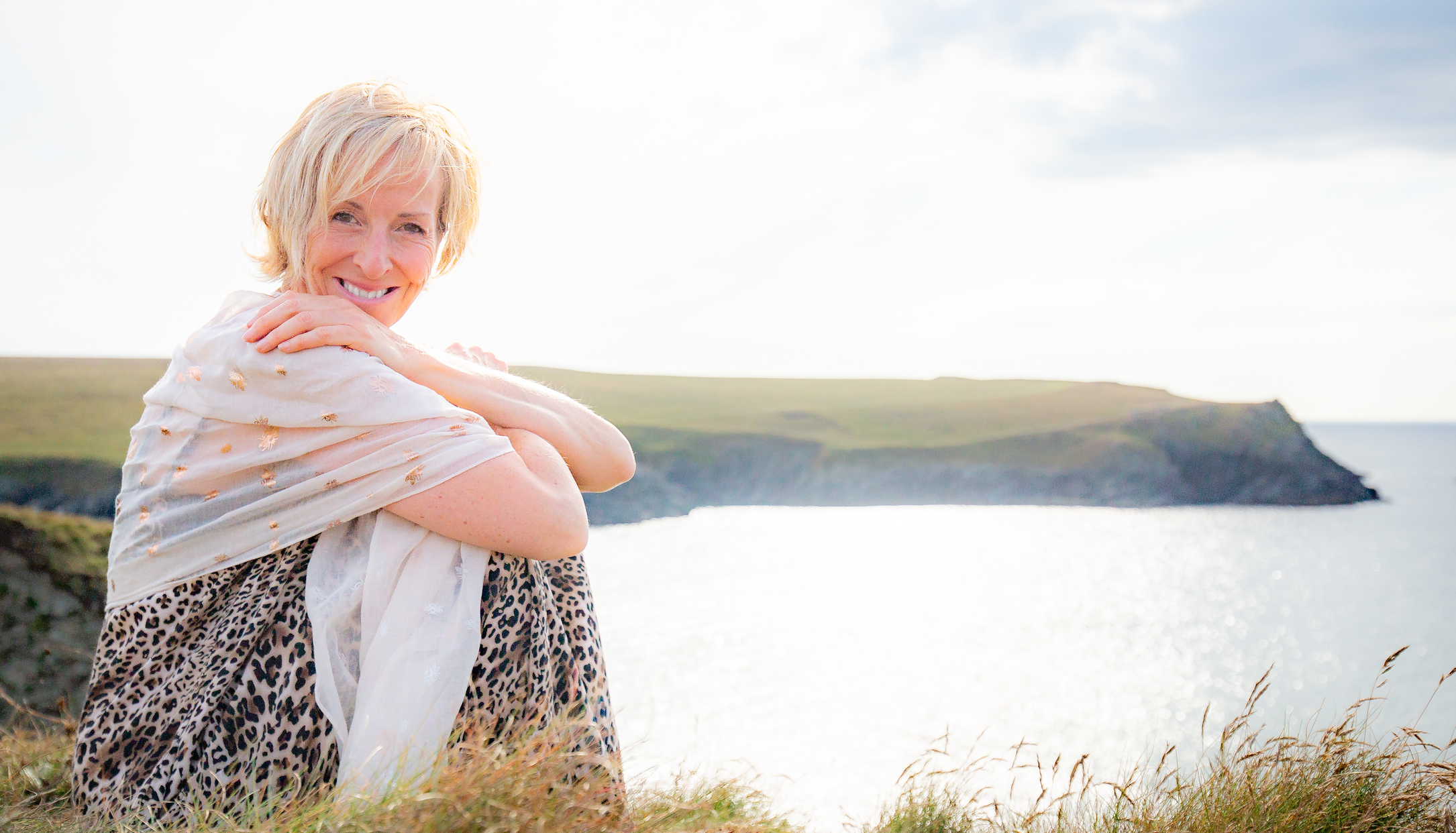 Julia Poole Artist sitting on Cornwall clifftop with sea sparkling in background