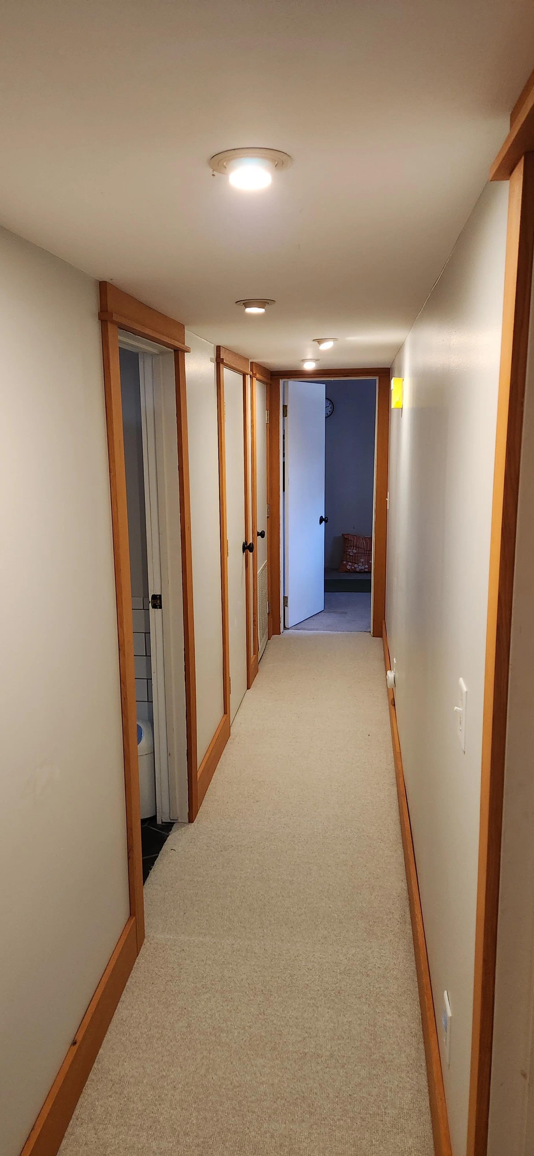 A narrow hallway with beige carpet, light-colored walls, and wooden trim. This picture showcases many header pieces of stain grade trim scribed to the ceiling. The casing legs at the end of the hallway are also scribed to the walls.