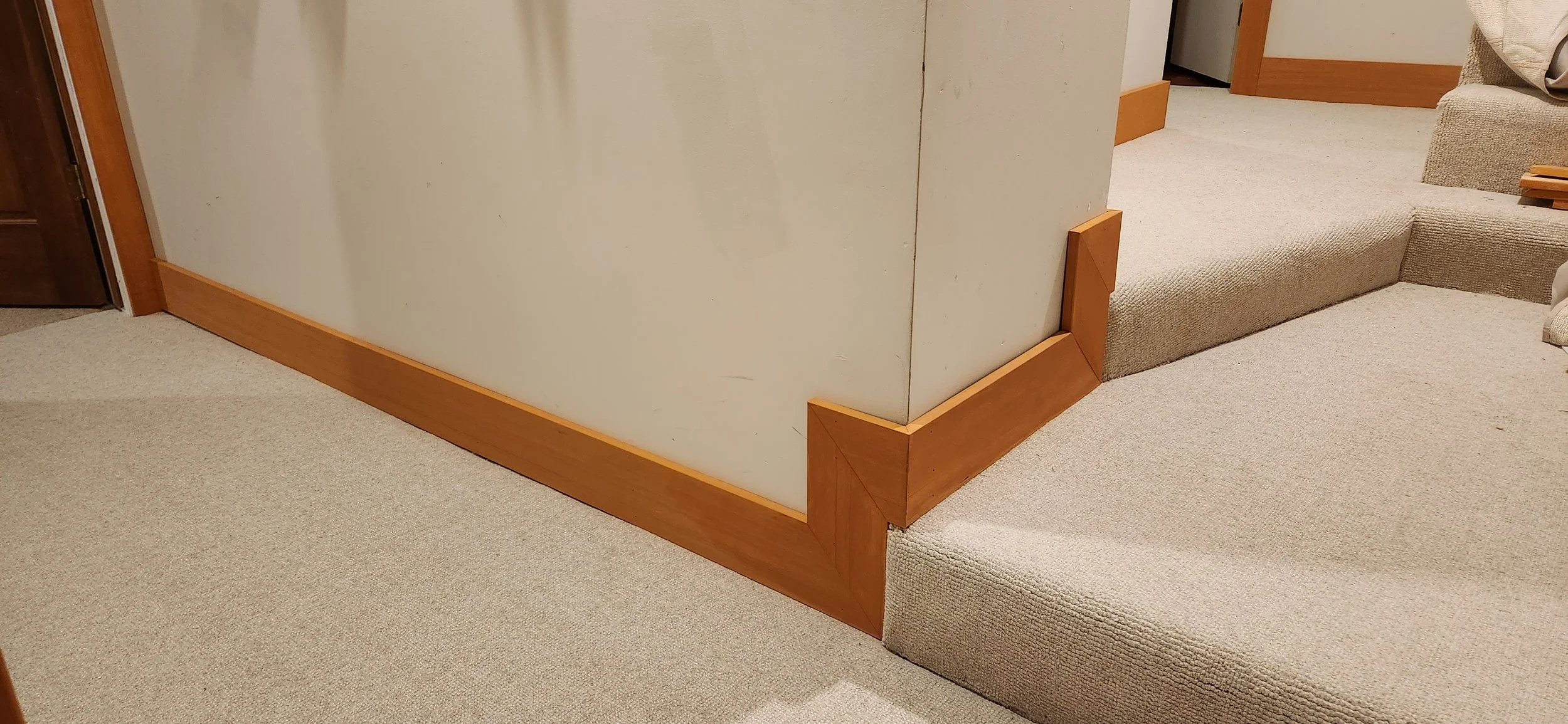 A hallway with beige carpet and light gray walls. Showcasing 5 inch hemlock stained trim wraping up along 2 landings.