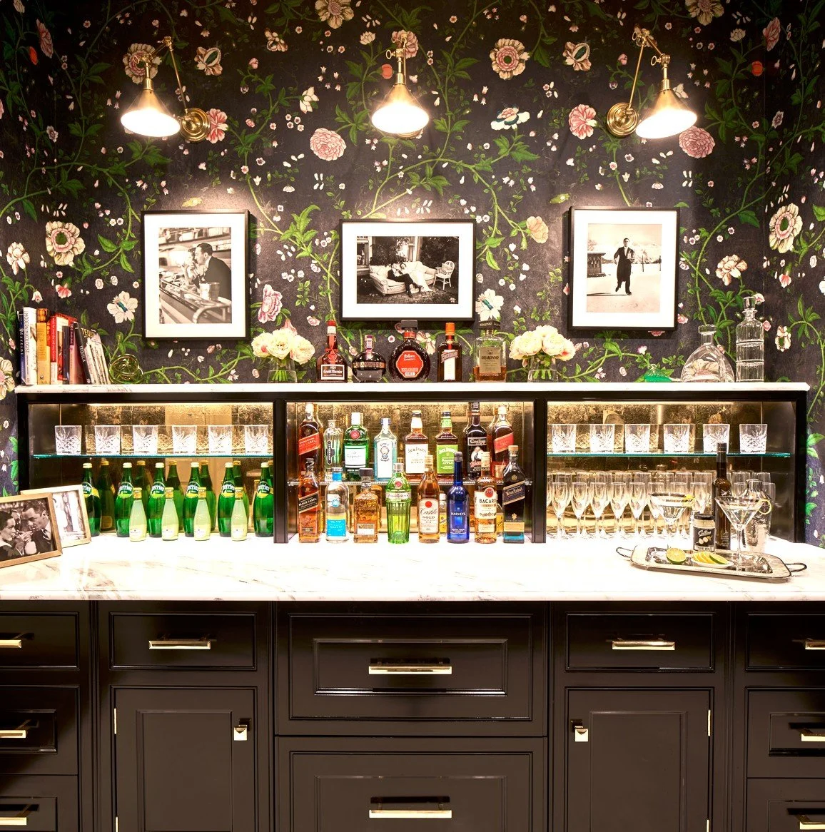 Home bar setup with black cabinetry, white marble countertop, and floral wallpaper background. The bar displays various liquor bottles, glassware, books, and decorative items, illuminated by three gold wall-mounted lamps.