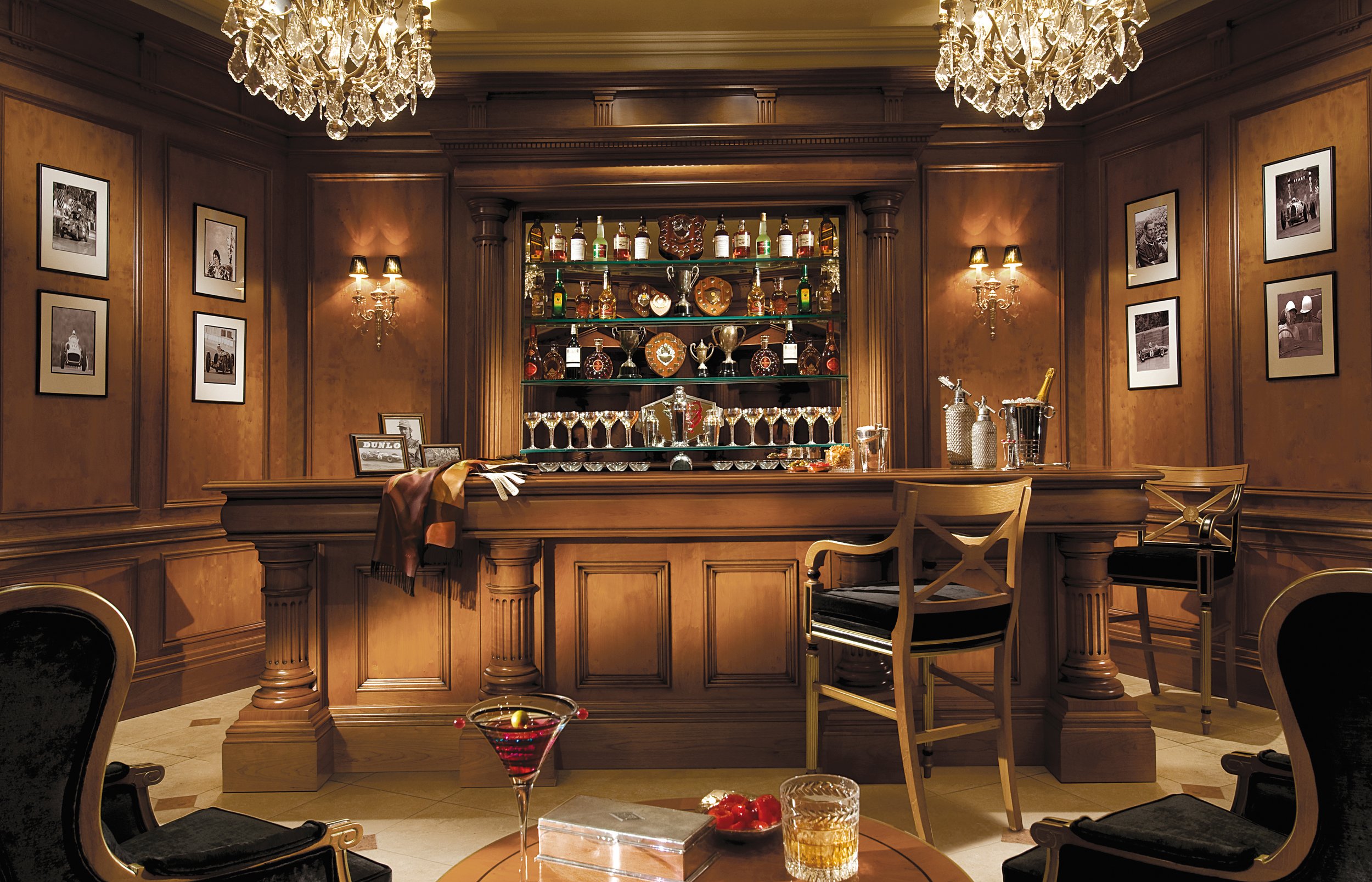 A luxurious home bar with wooden paneling, a glass shelving unit filled with liquor bottles, trophies, and glassware, two chandeliers, framed black-and-white photographs on the walls, and a round table with drinks and snacks in the foreground.